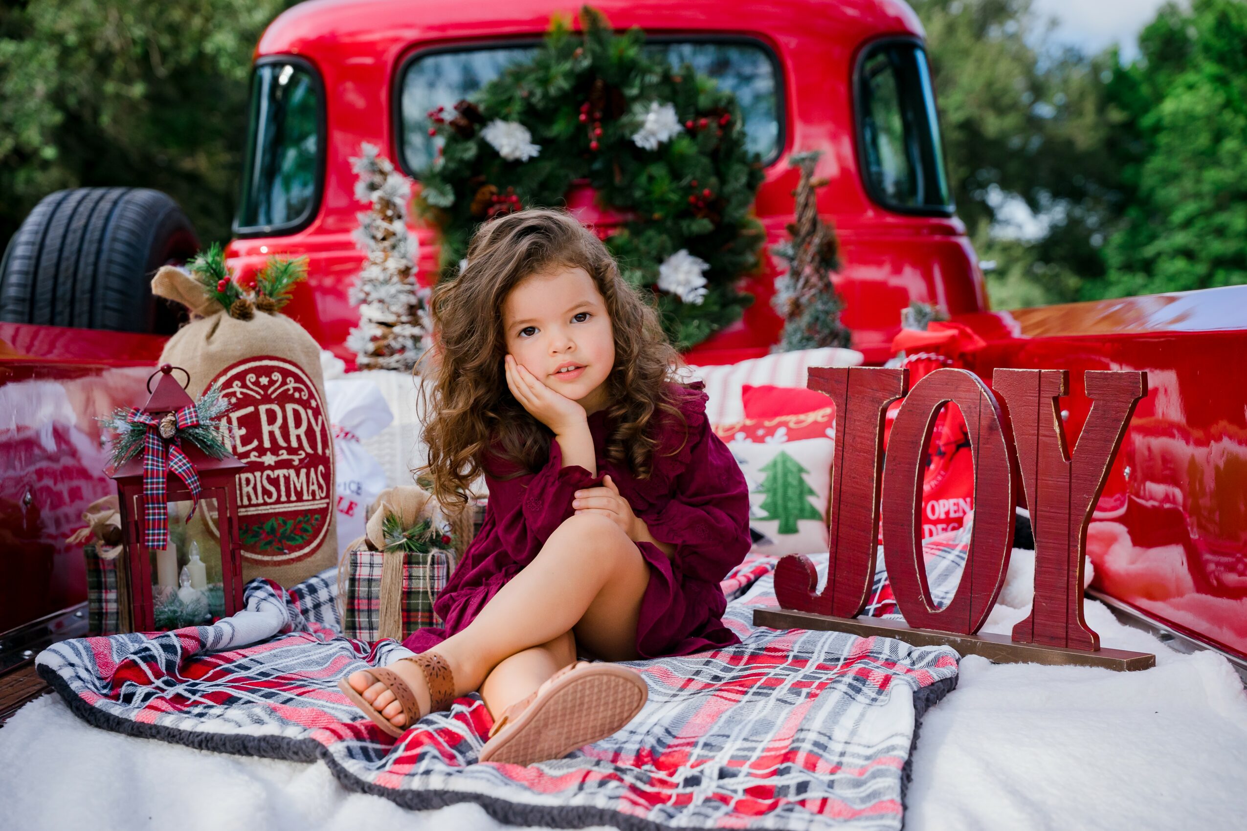 Young girl sitting in the back of a decorated red truck with Christmas ornaments and a large 'JOY' sign.
