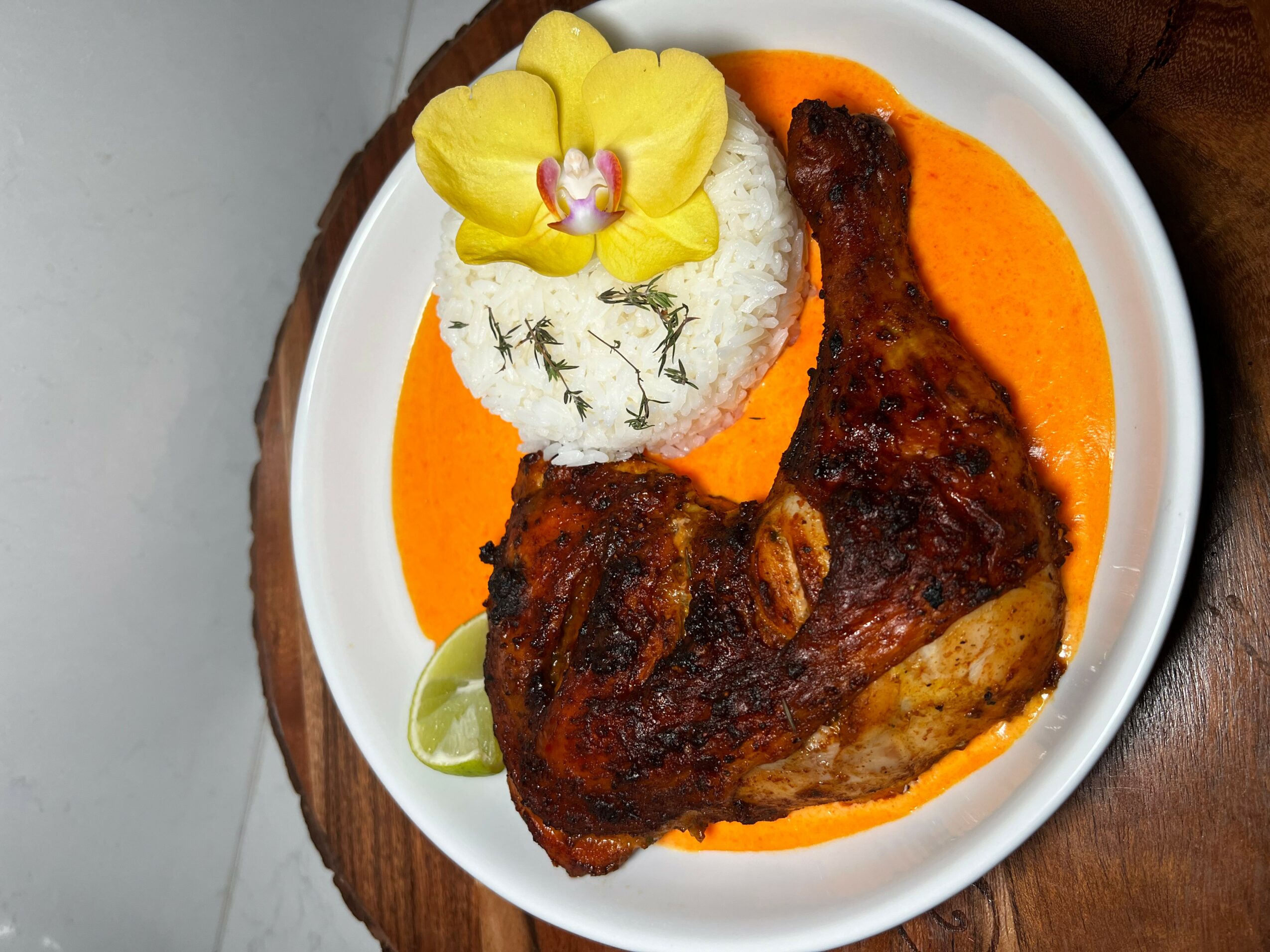 Plate with grilled chicken leg, rice topped with a yellow flower, and orange sauce, on a wooden surface.