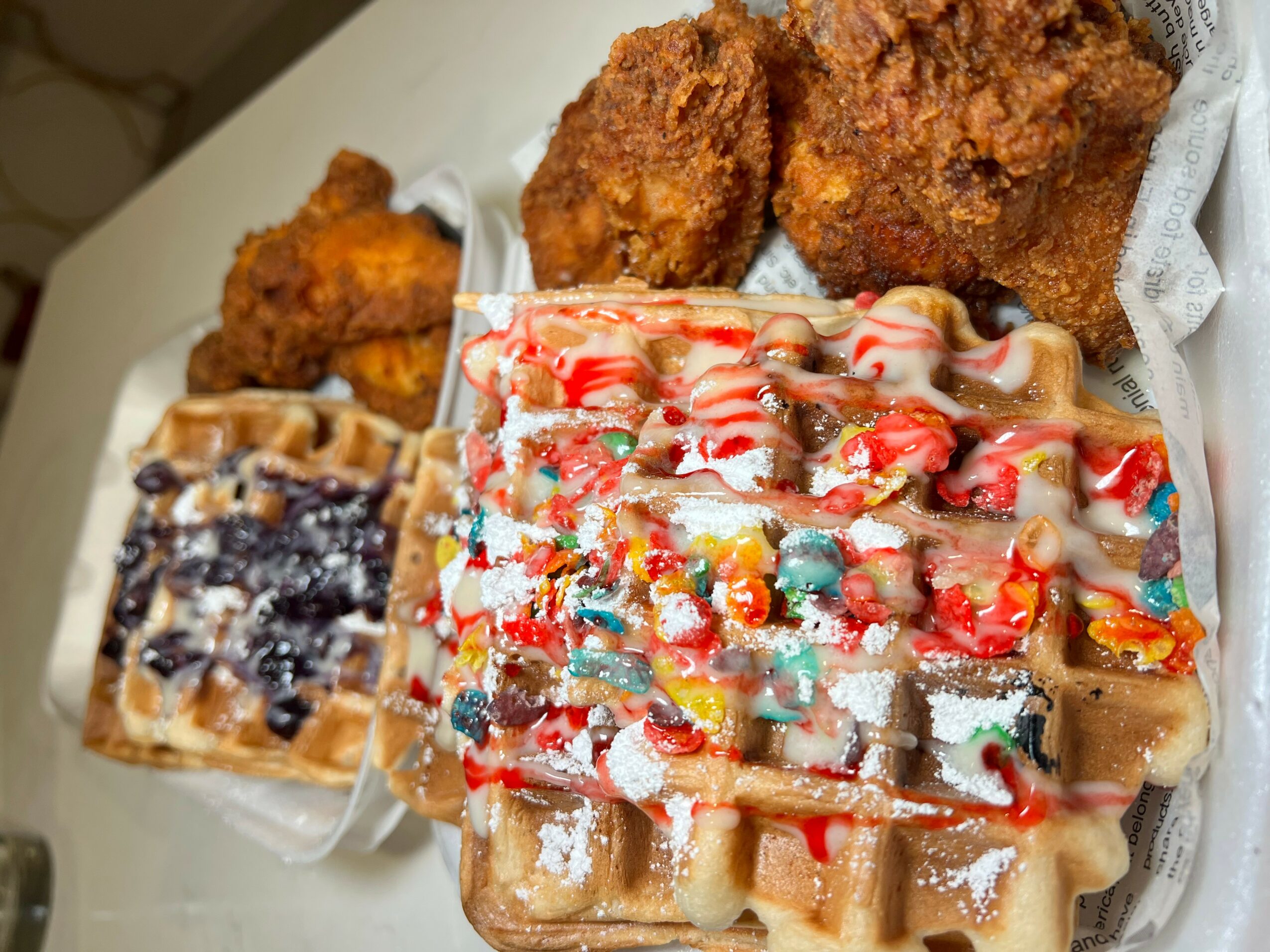Two waffles topped with colorful candies and drizzled with caramel, alongside fried chicken pieces on a white surface.