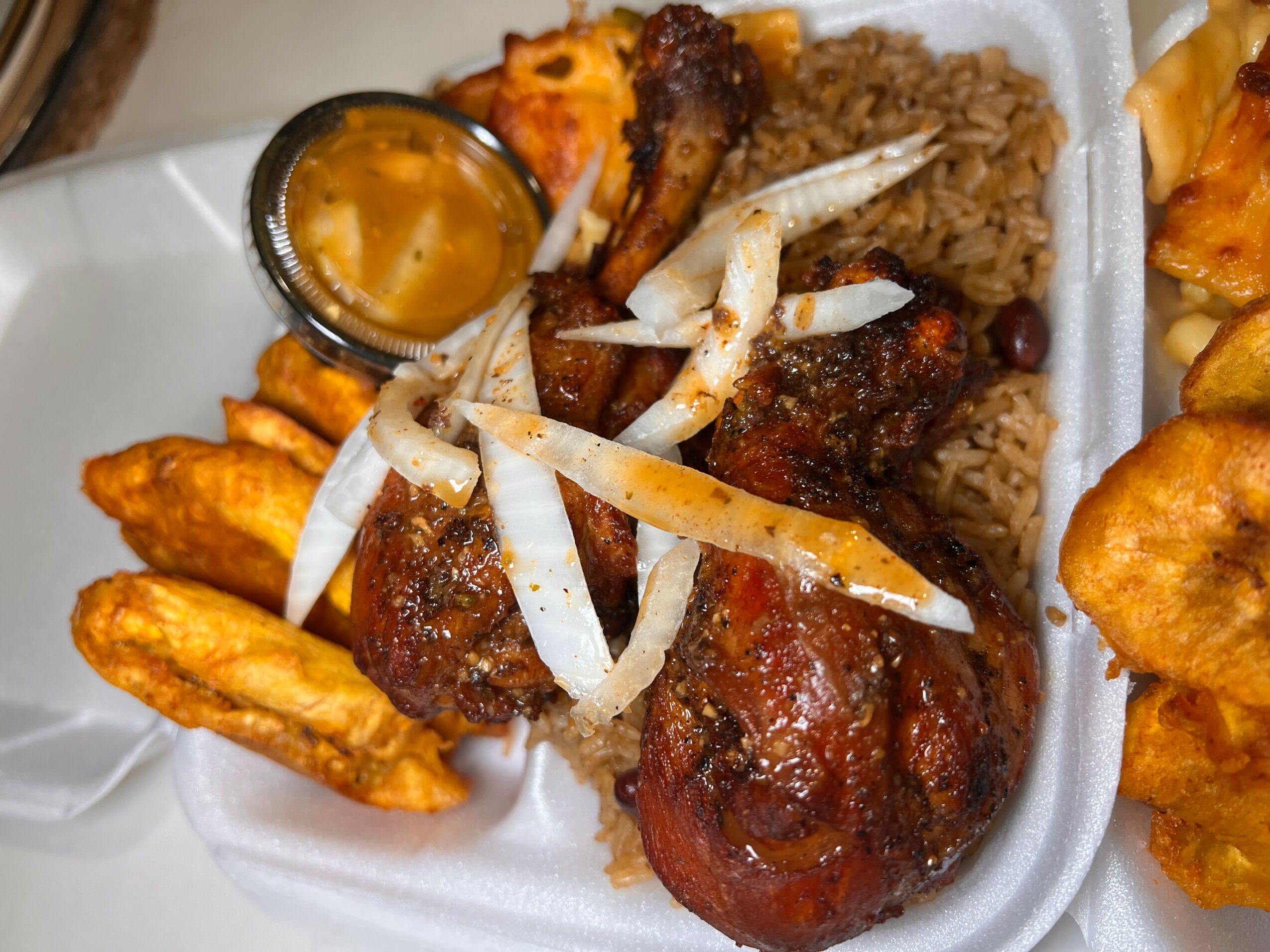 Fried chicken, rice, fried plantains, and a small container of sauce on a foam plate.