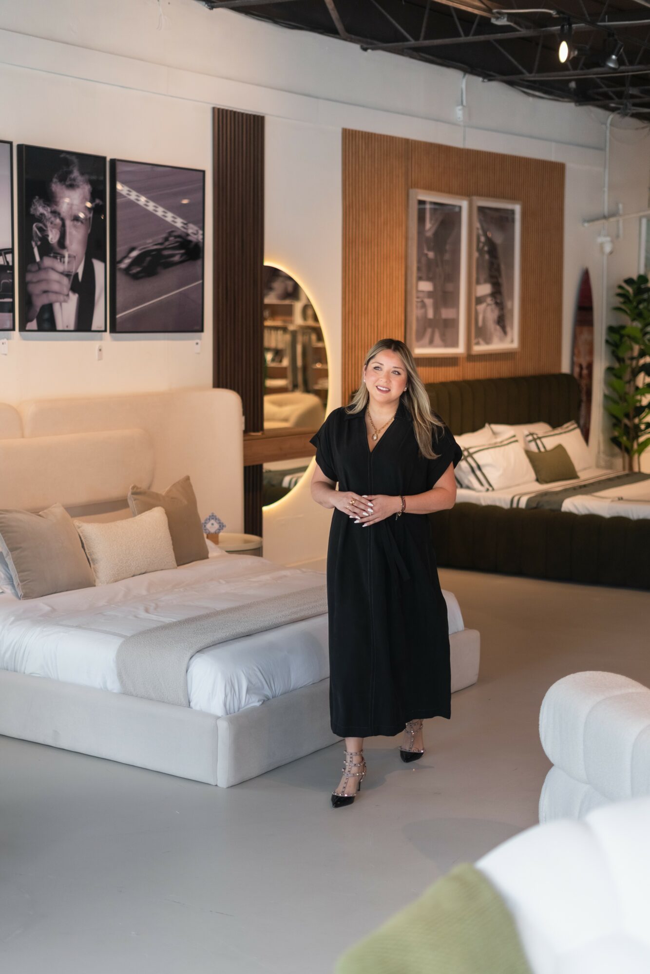 Woman in black dress standing in a furniture store with beds and artwork on the wall behind her.