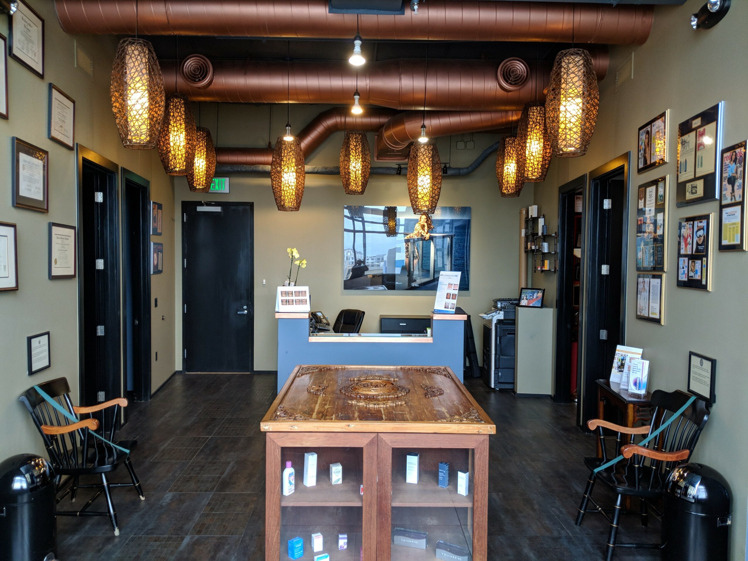Reception area with a wooden table, framed pictures on walls, chairs, and decorative hanging lights, with a window behind the desk.