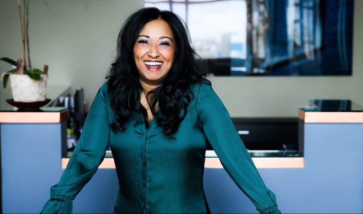 Woman with long dark hair smiling, wearing a teal blouse, standing in an office with blue and white decor.