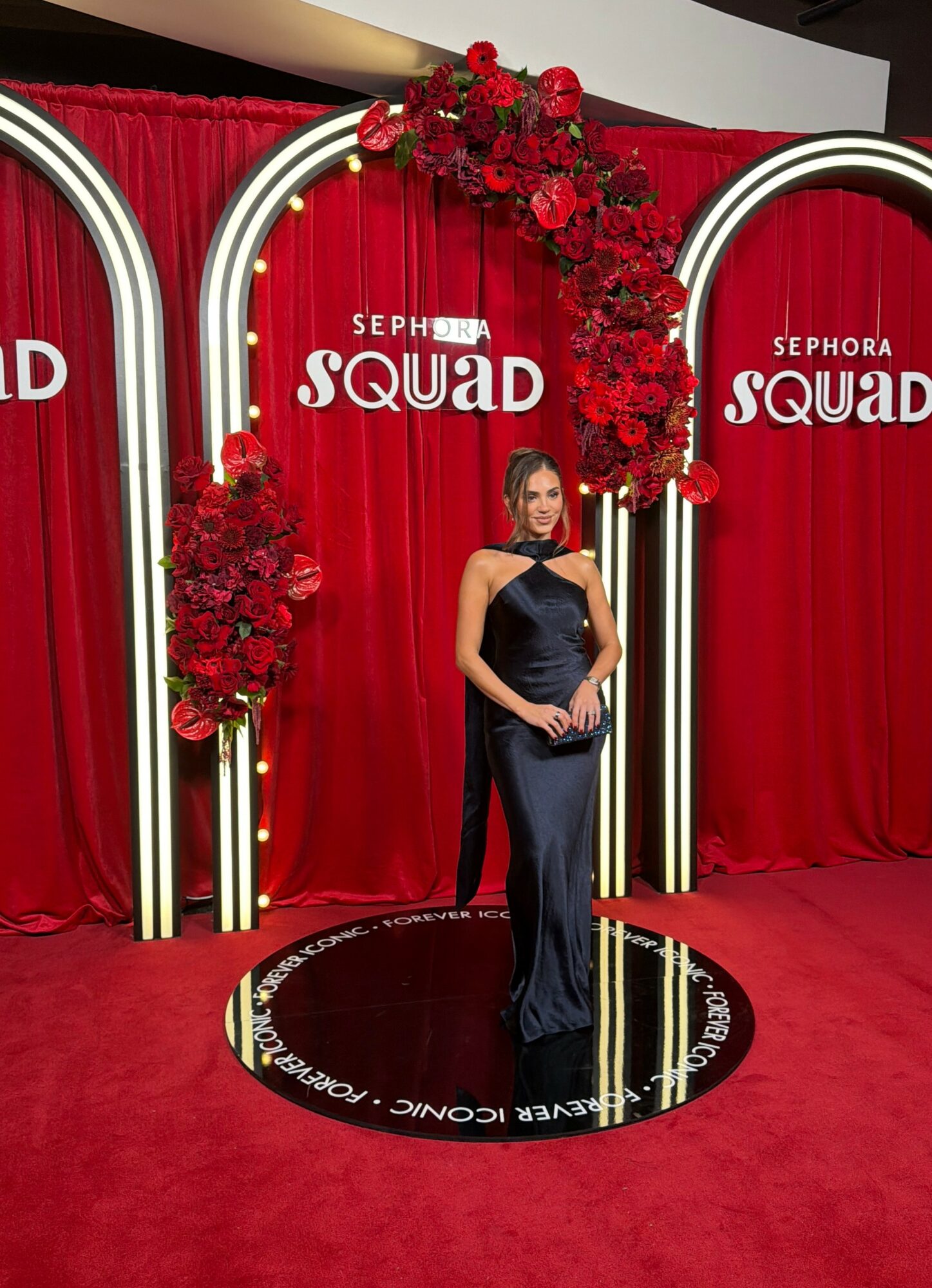 Woman in a dark gown standing on a circular platform with red curtains and floral decorations behind her.