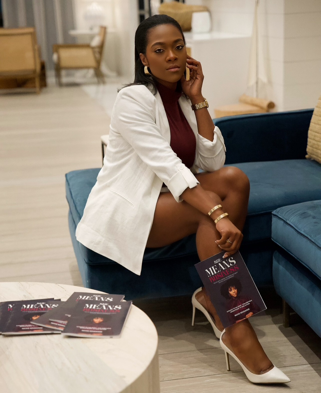 Woman sitting on a blue sofa, wearing a white blazer, maroon dress, and high heels, holding a book, in a room with light-colored flooring and furniture.