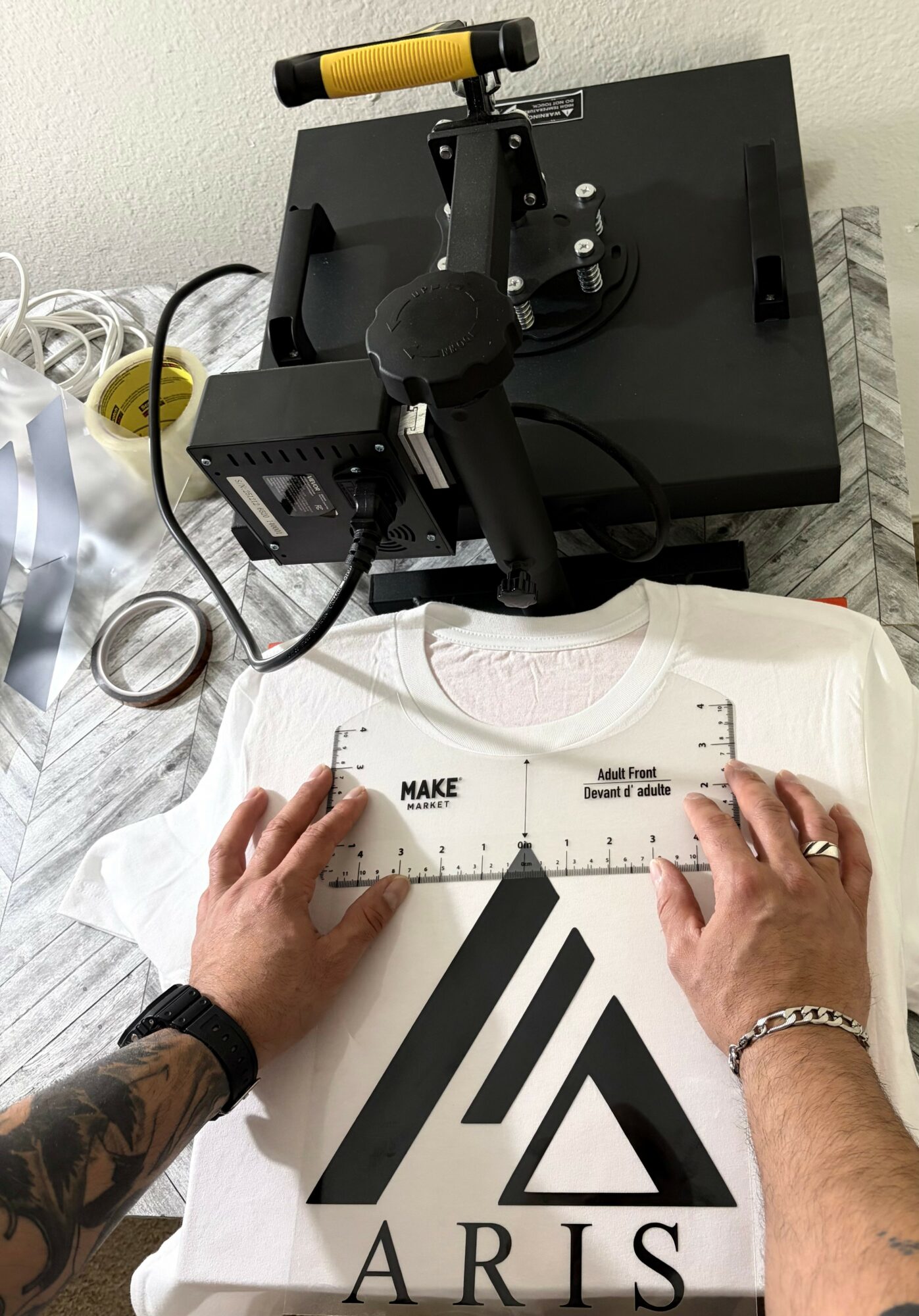 Person using a ruler on a white shirt with a black logo, with a black device and wires nearby on a surface.