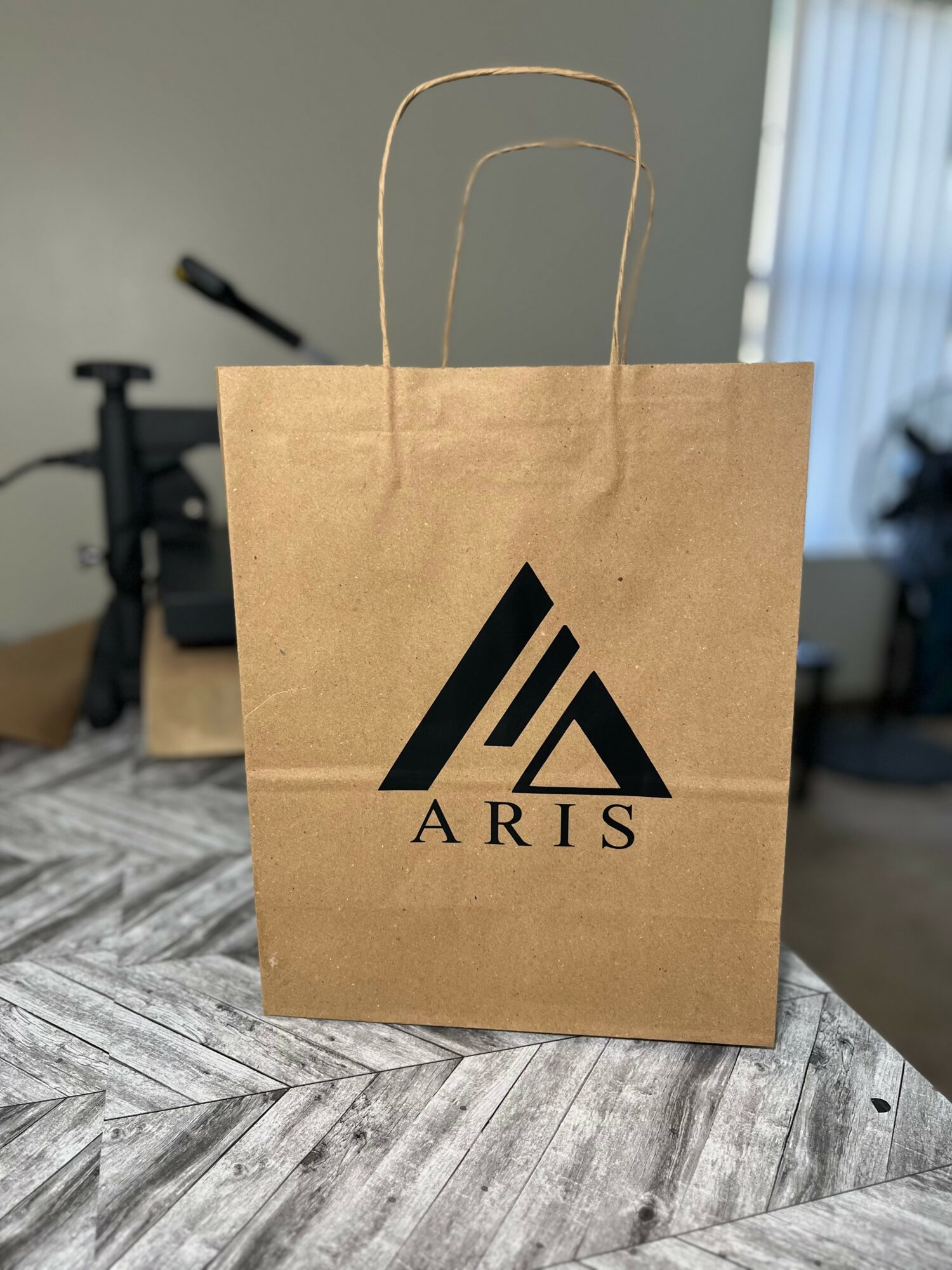 Brown paper shopping bag with black logo and handles on a wooden table, background shows a room with a window and equipment.