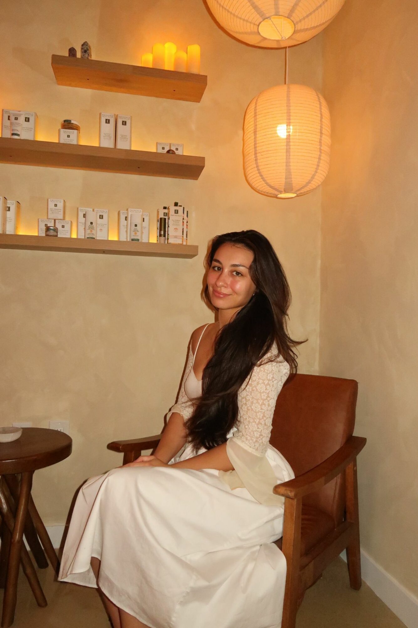 Woman with long dark hair sitting on a wooden chair in a cozy room with warm lighting and decorative shelves.
