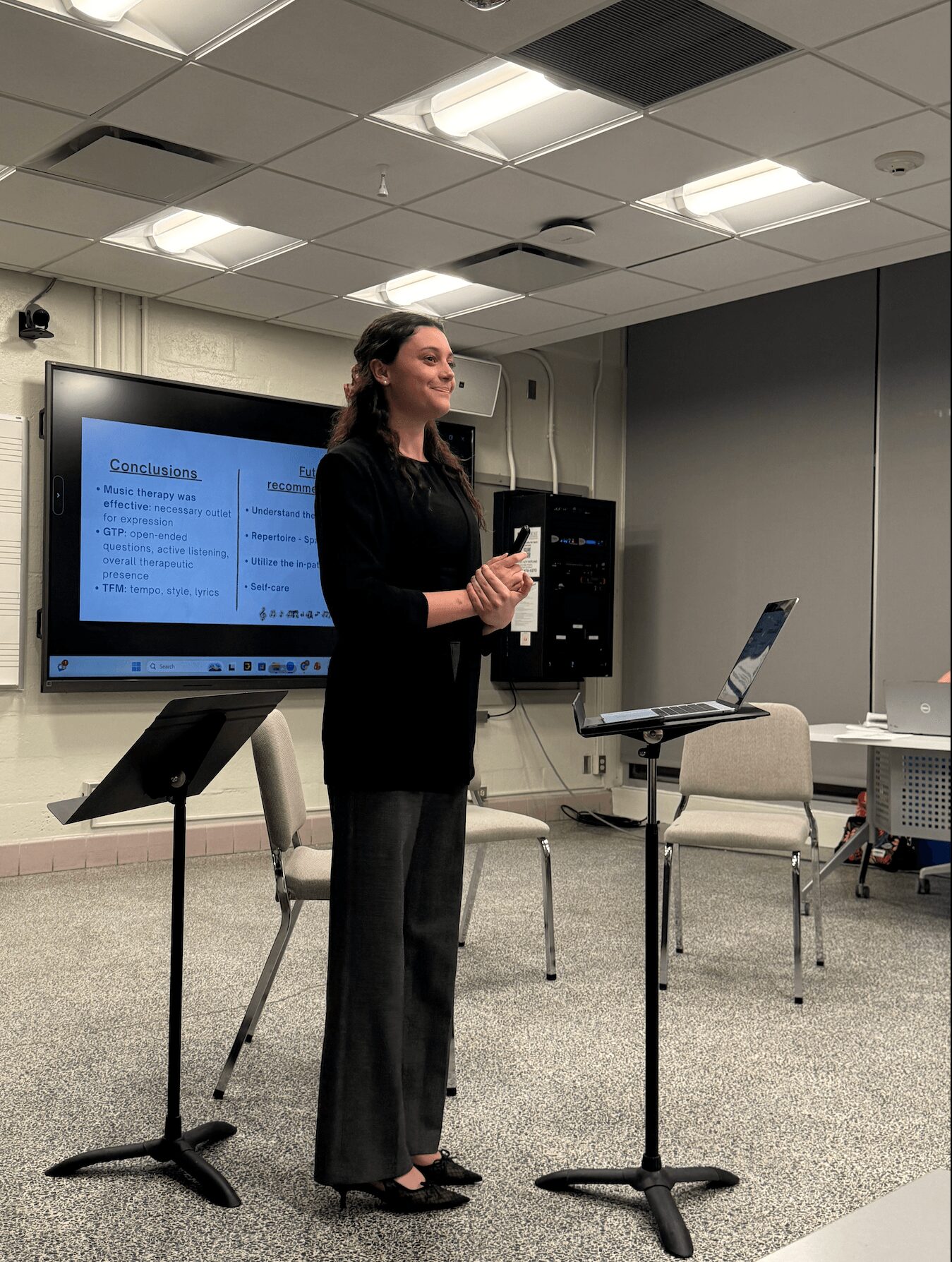 Woman standing in a conference room, holding a phone, with a presentation slide on a screen behind her.