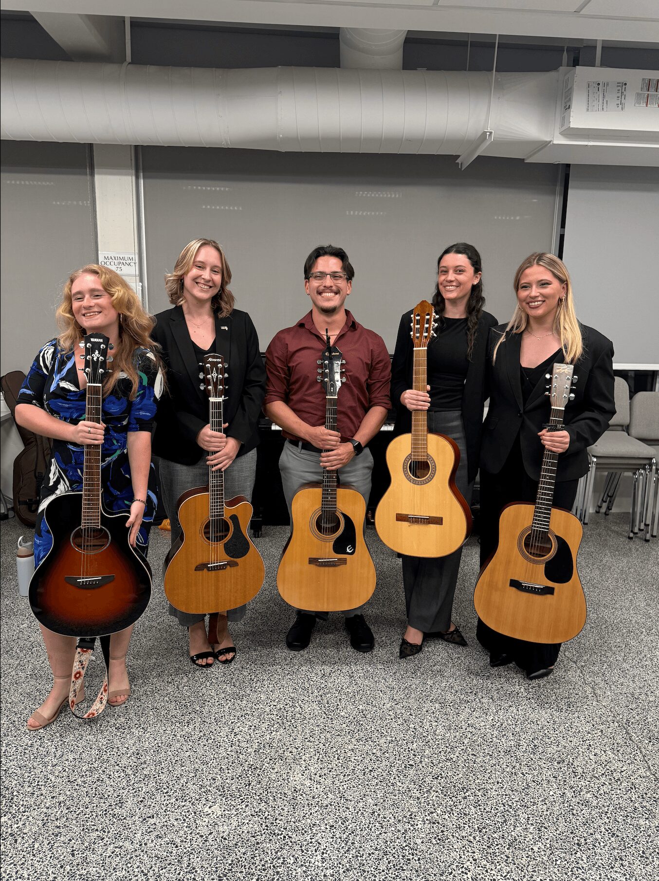 Five people holding guitars, standing indoors against a plain wall, smiling, with a gray carpeted floor.