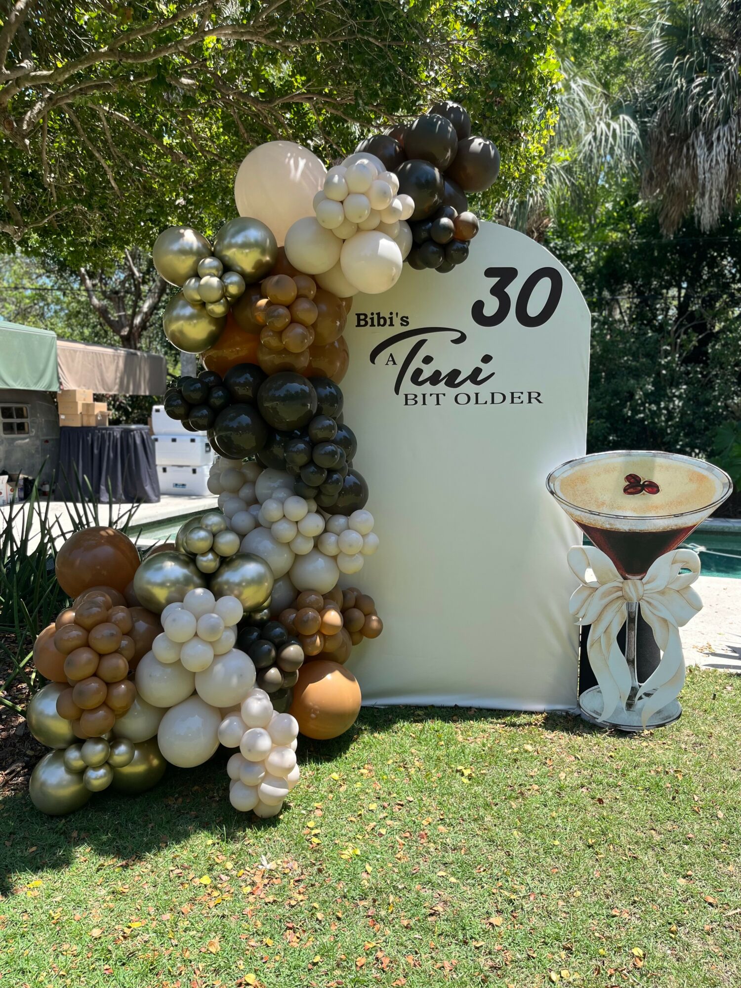 Balloon arrangement with white, black, gold, and brown balloons next to a white sign with black text, outdoors on grass.