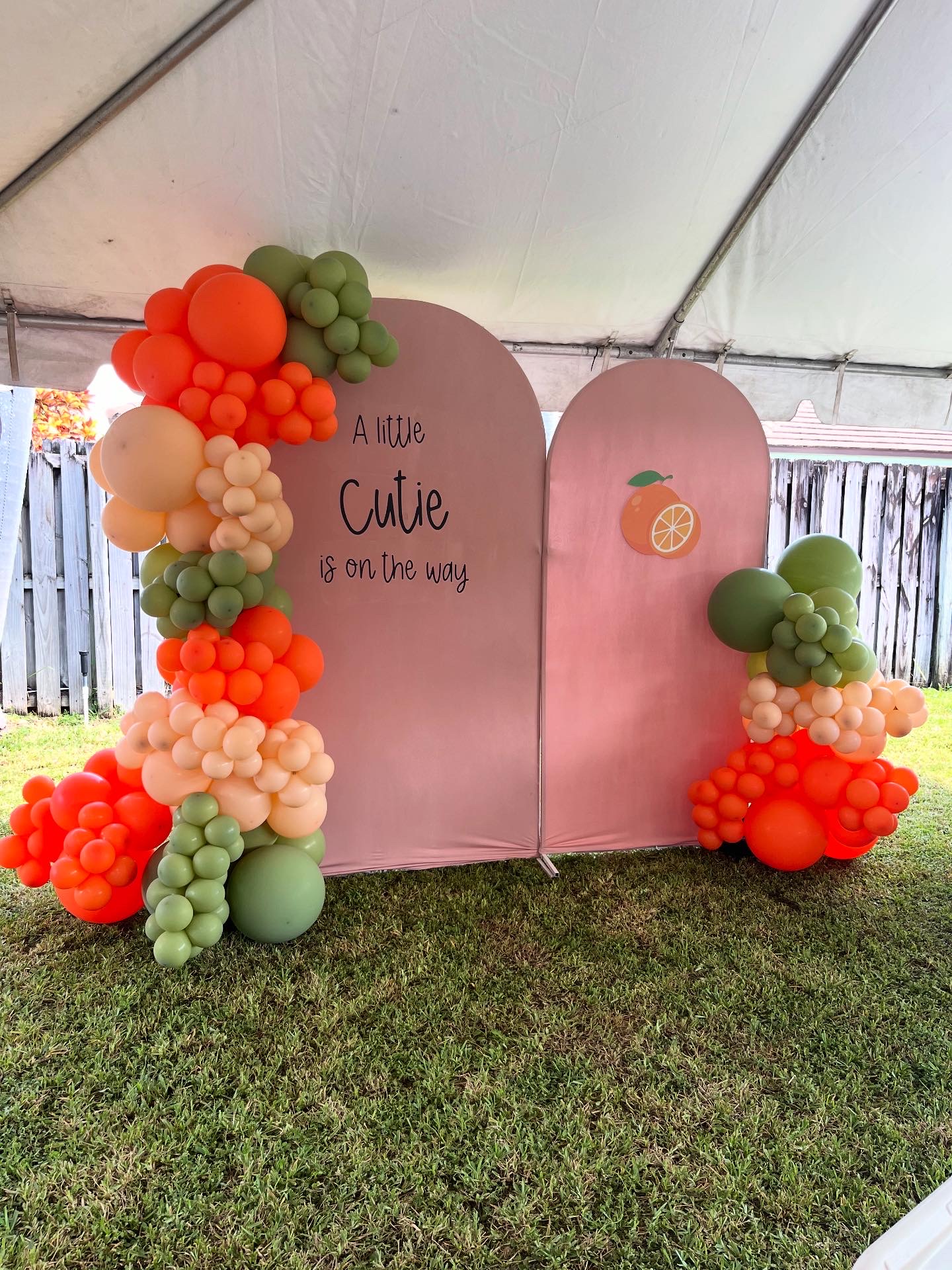 Decorative backdrop with pink panels and balloon arrangements in orange, green, and cream, with a handwritten sign, outdoors on grass.