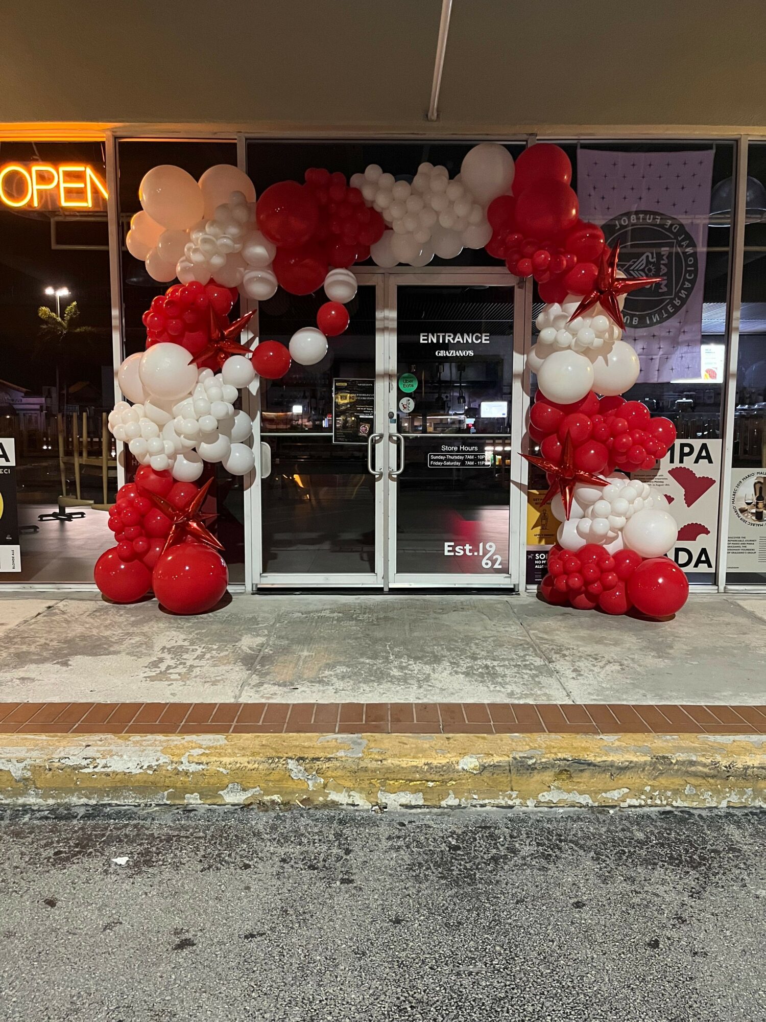 Store window decorated with red and white balloons, with a neon sign reading 'OPEN' and a street view outside.