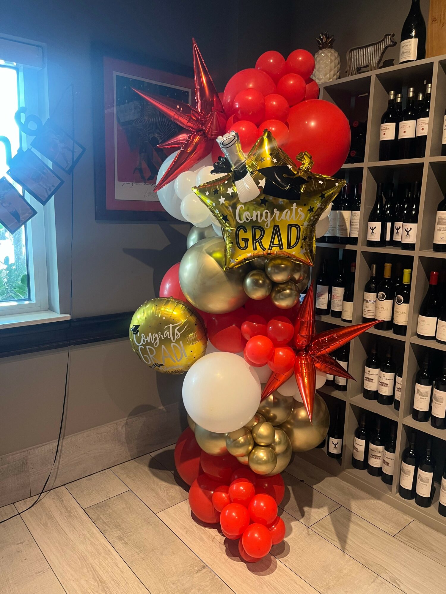 Arrangement of red, gold, and white balloons with star-shaped balloons and a gold 'Graduation' balloon, on a wooden floor.