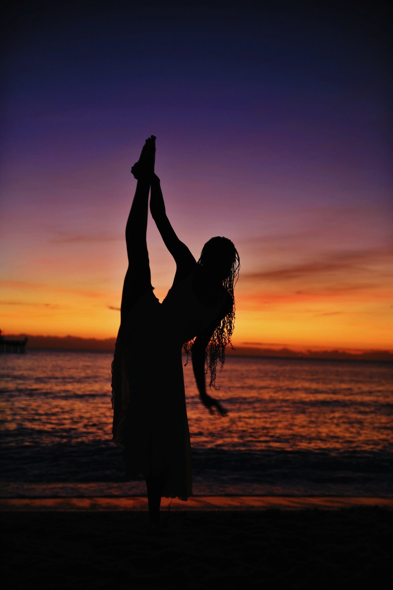 Silhouette of a person performing a yoga pose on the beach during sunset, with colorful sky and water in background.