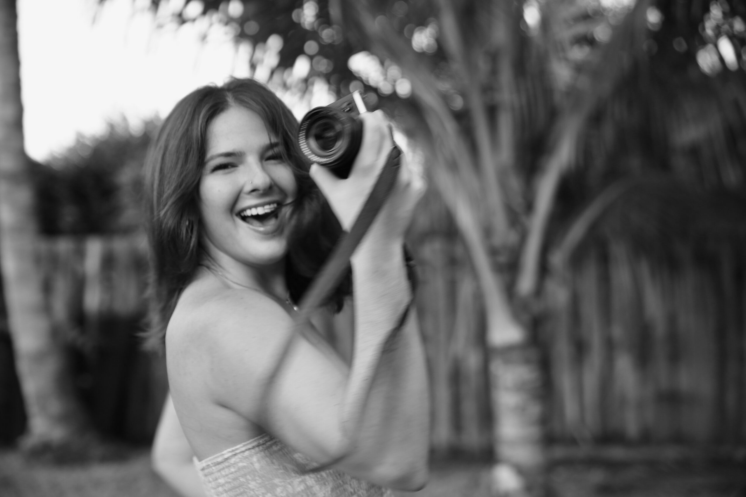 Woman smiling and holding a camera outdoors, with trees and a fence in the background.