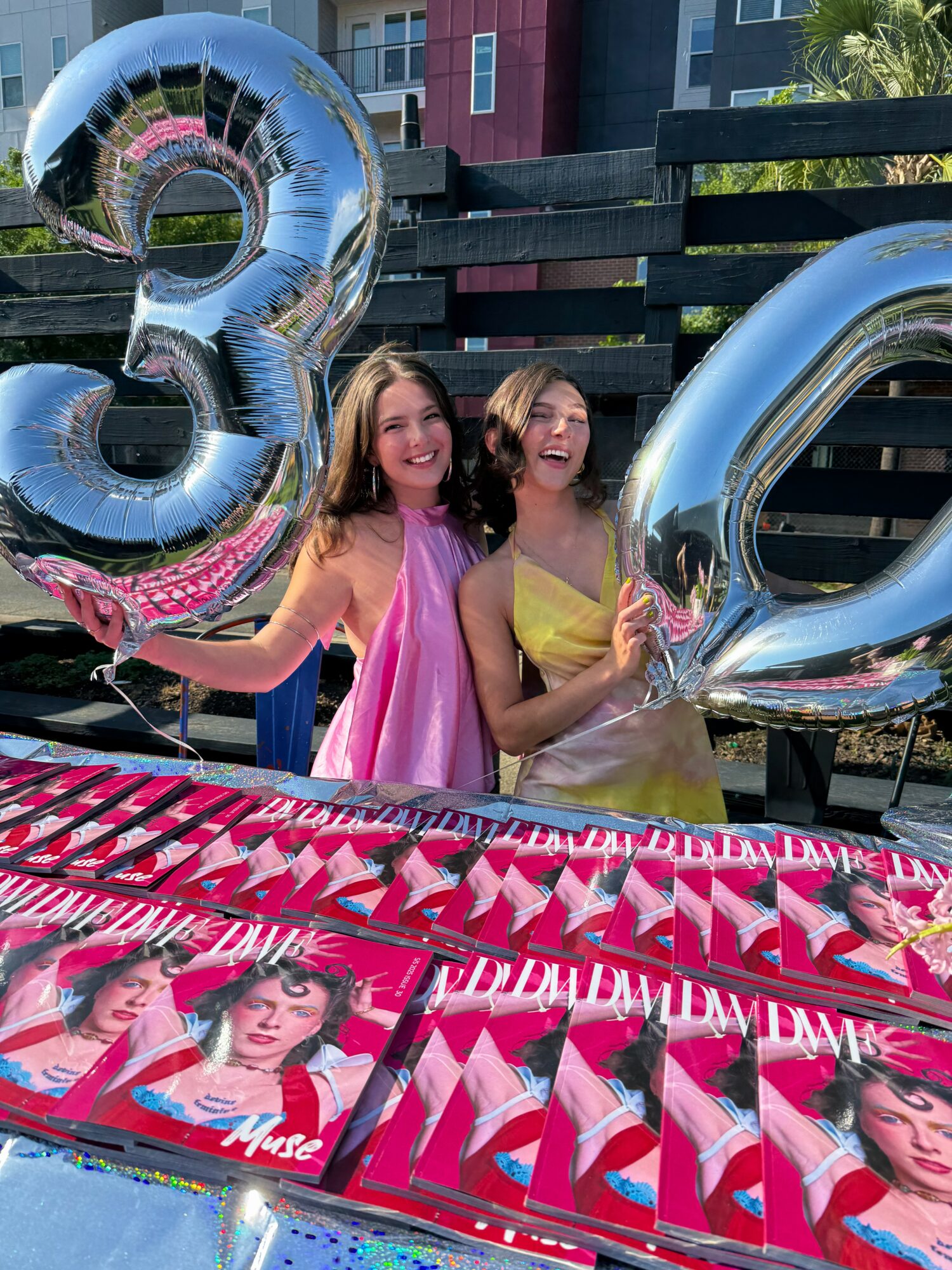 Two young women holding large silver balloons shaped as the numbers 3 and 2, smiling outdoors with a table of magazines.