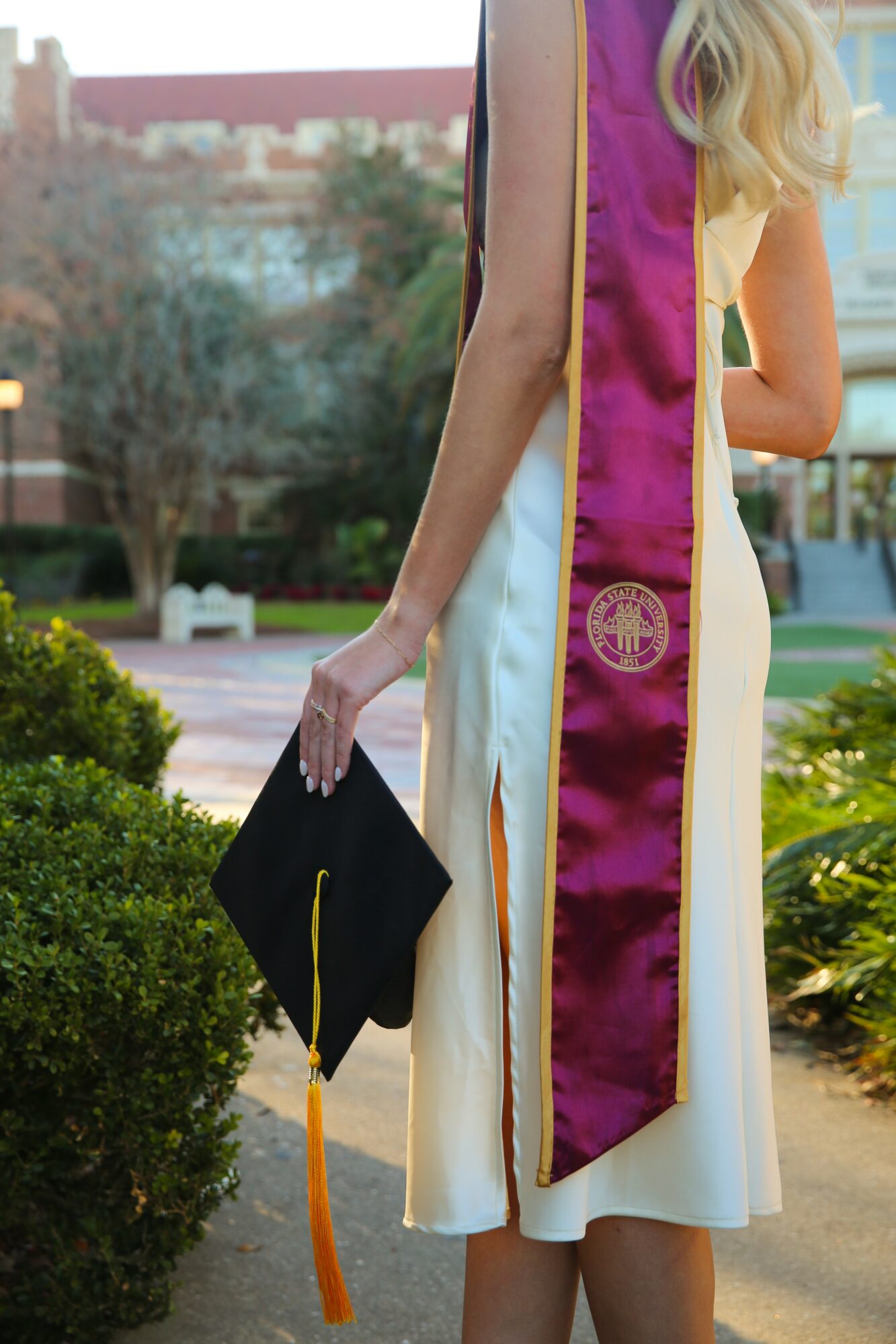 Person in graduation gown holding a cap, standing outdoors with trees and buildings in background.