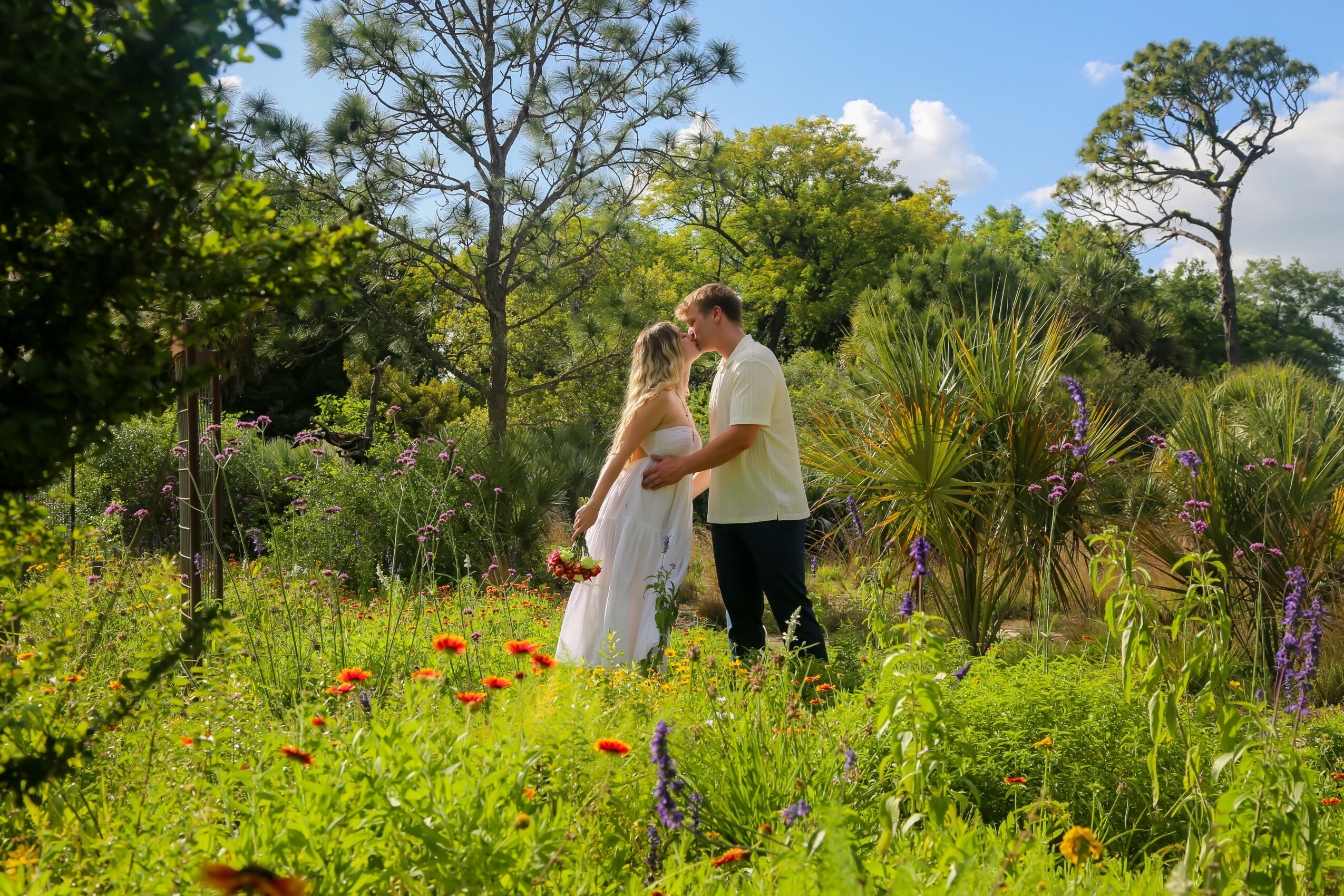 A couple stands close together in a lush garden with trees and colorful flowers, sharing a moment.