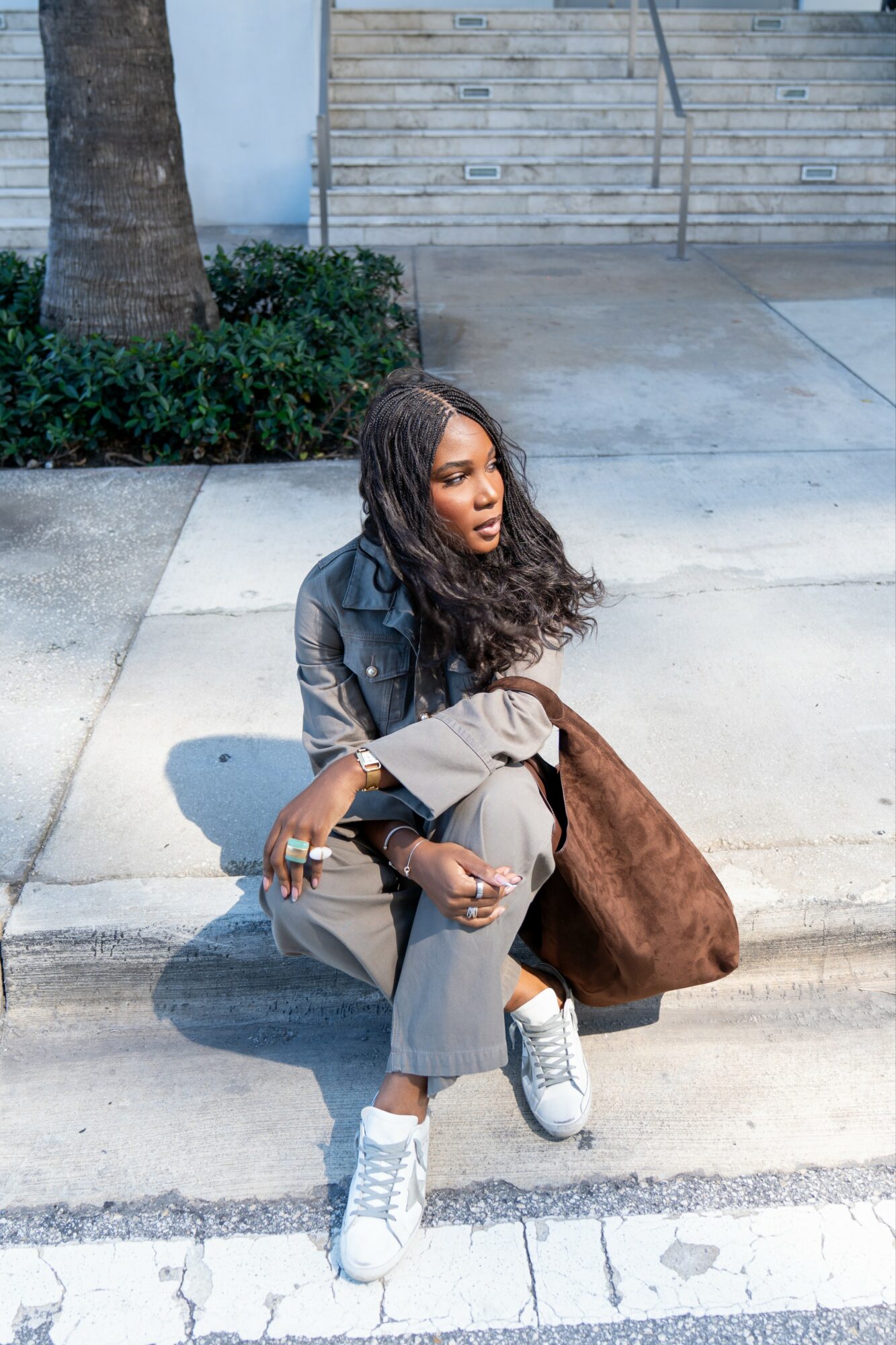 Young woman sitting on sidewalk with a large brown bag, wearing a leather jacket and sneakers, near stairs and a tree.