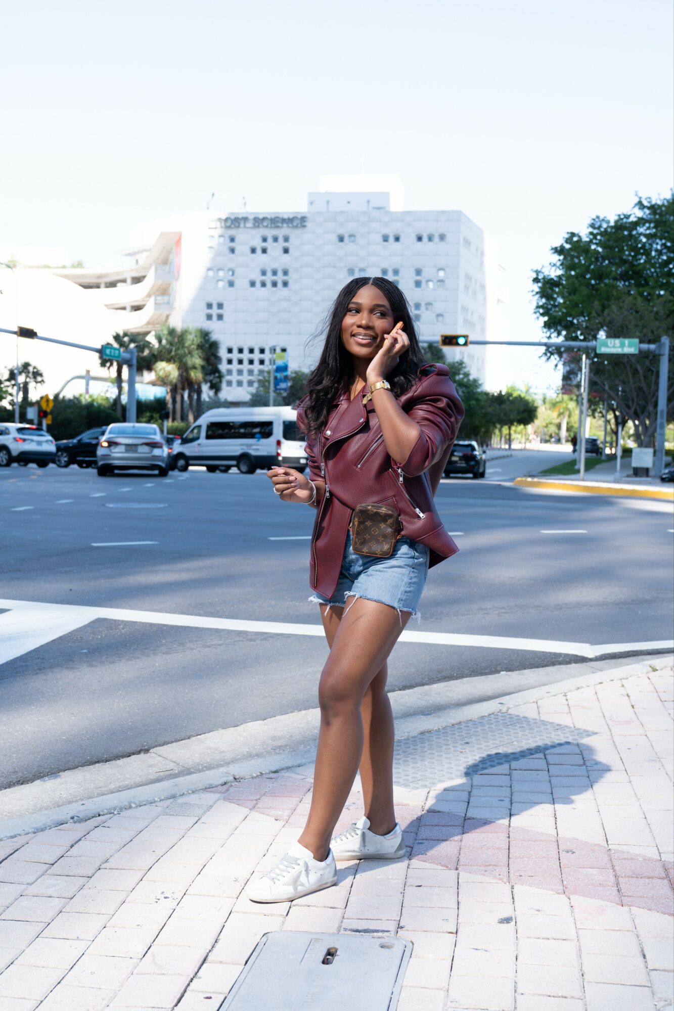 Young woman walking on sidewalk, talking on phone, wearing leather jacket, shorts, and sneakers, with city buildings and trees in background.