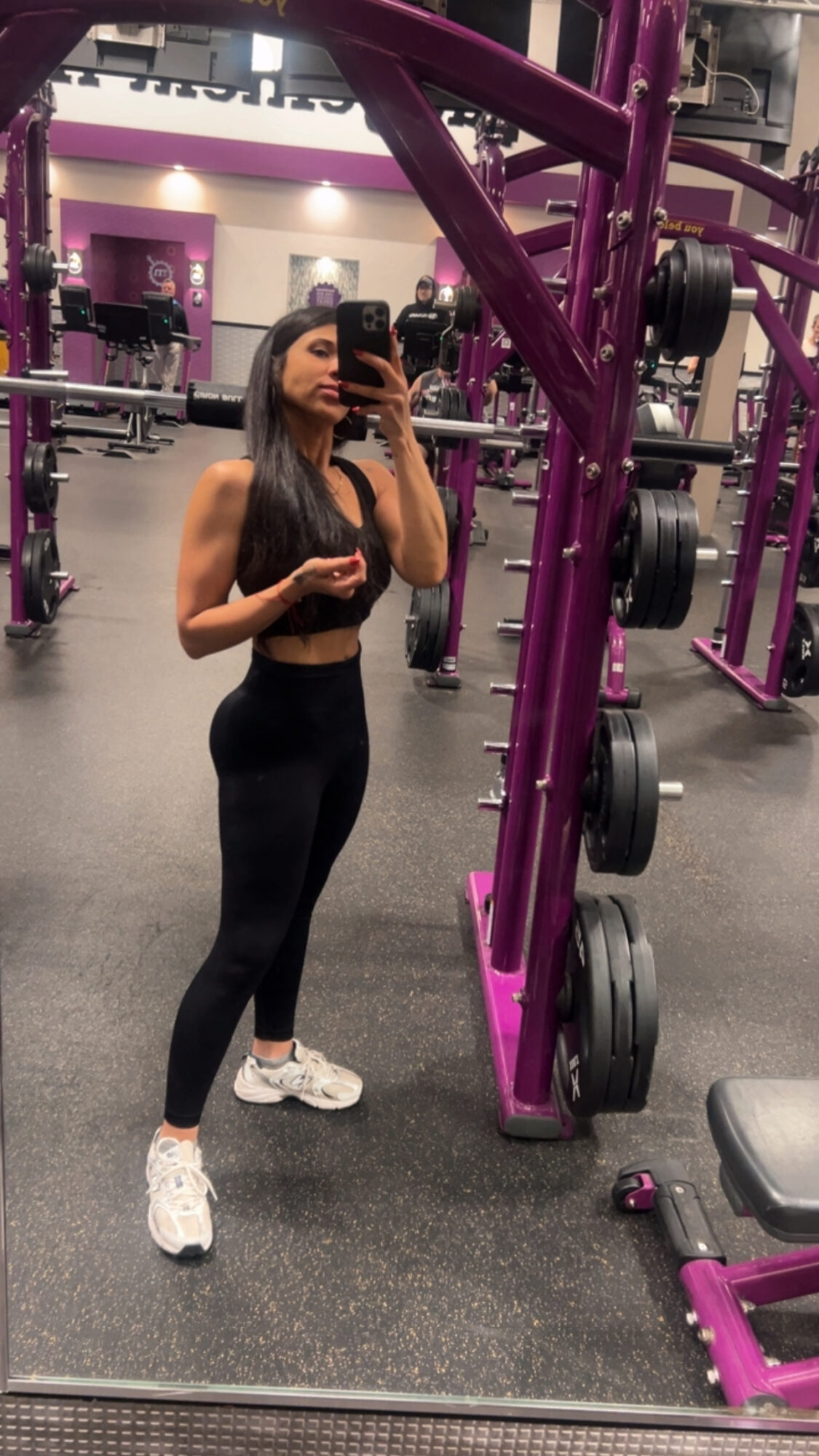 Woman taking mirror selfie in gym, wearing black workout clothes, purple gym equipment visible behind her.