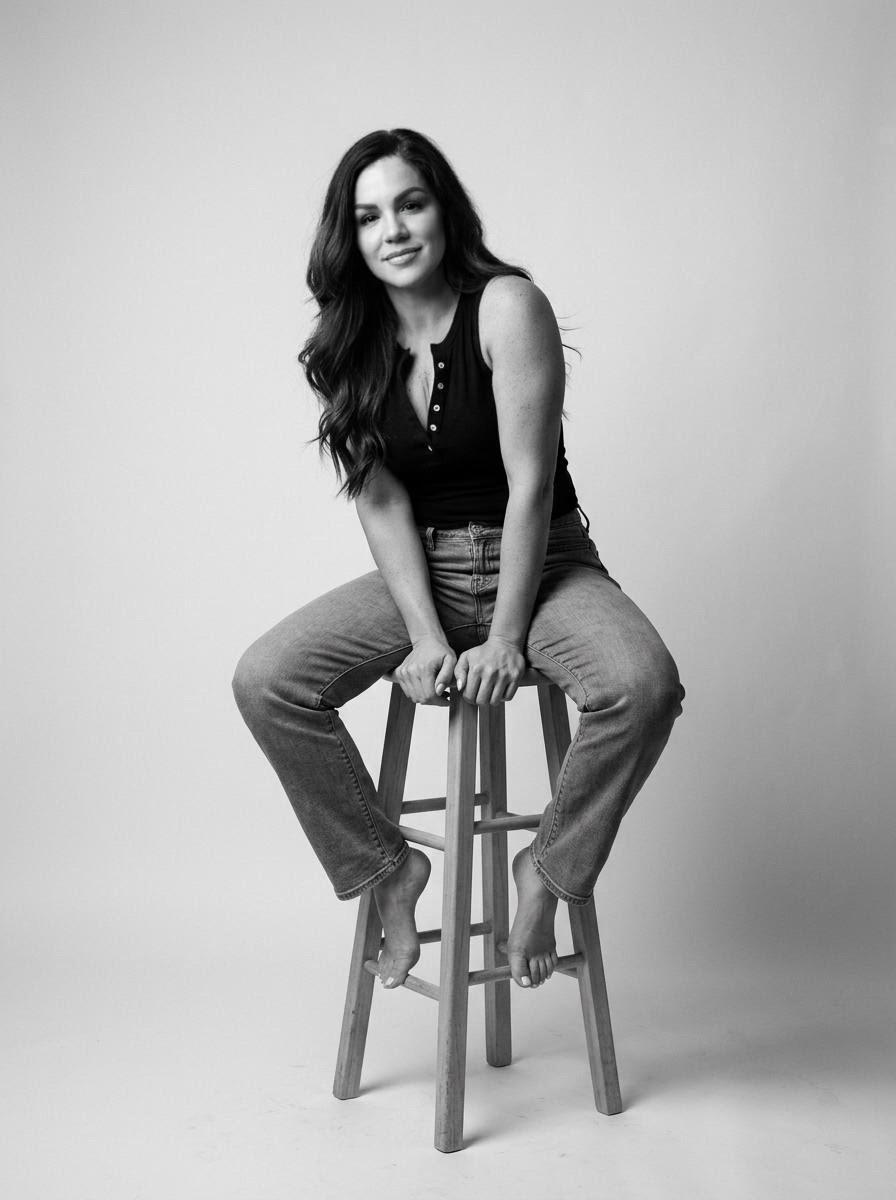 Woman sitting on a stool, smiling, with long wavy hair, wearing a sleeveless top and jeans, in a studio setting.