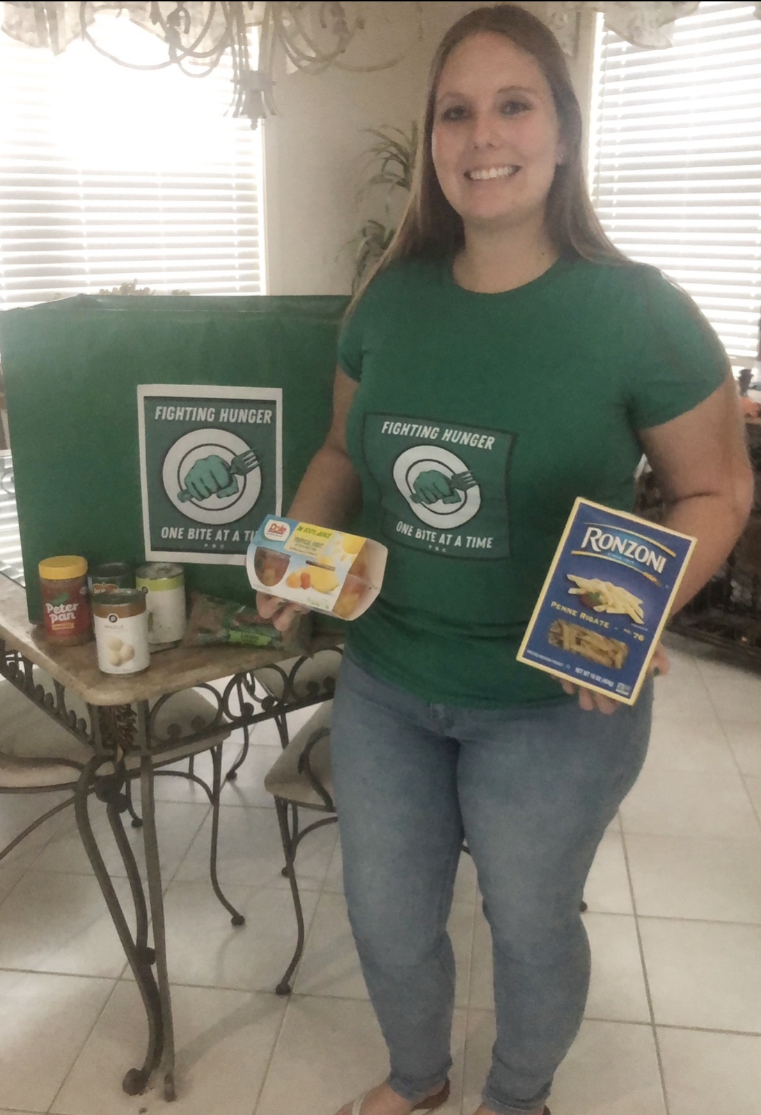 Woman holding food items in front of a table with donation boxes, smiling, indoors with windows and blinds behind her.