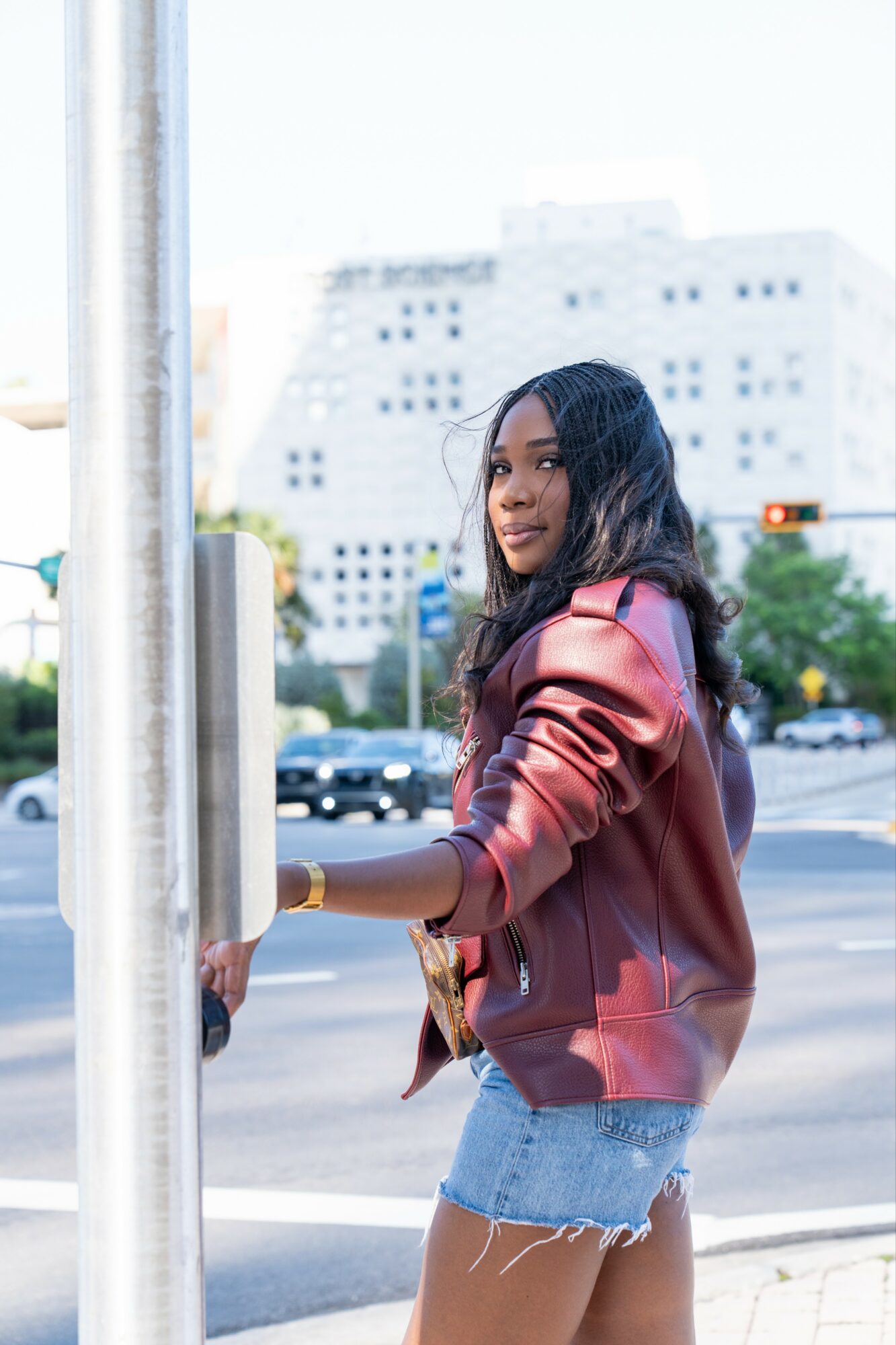 Young woman with long dark hair wearing a maroon jacket and denim shorts standing near a pole on a city street.