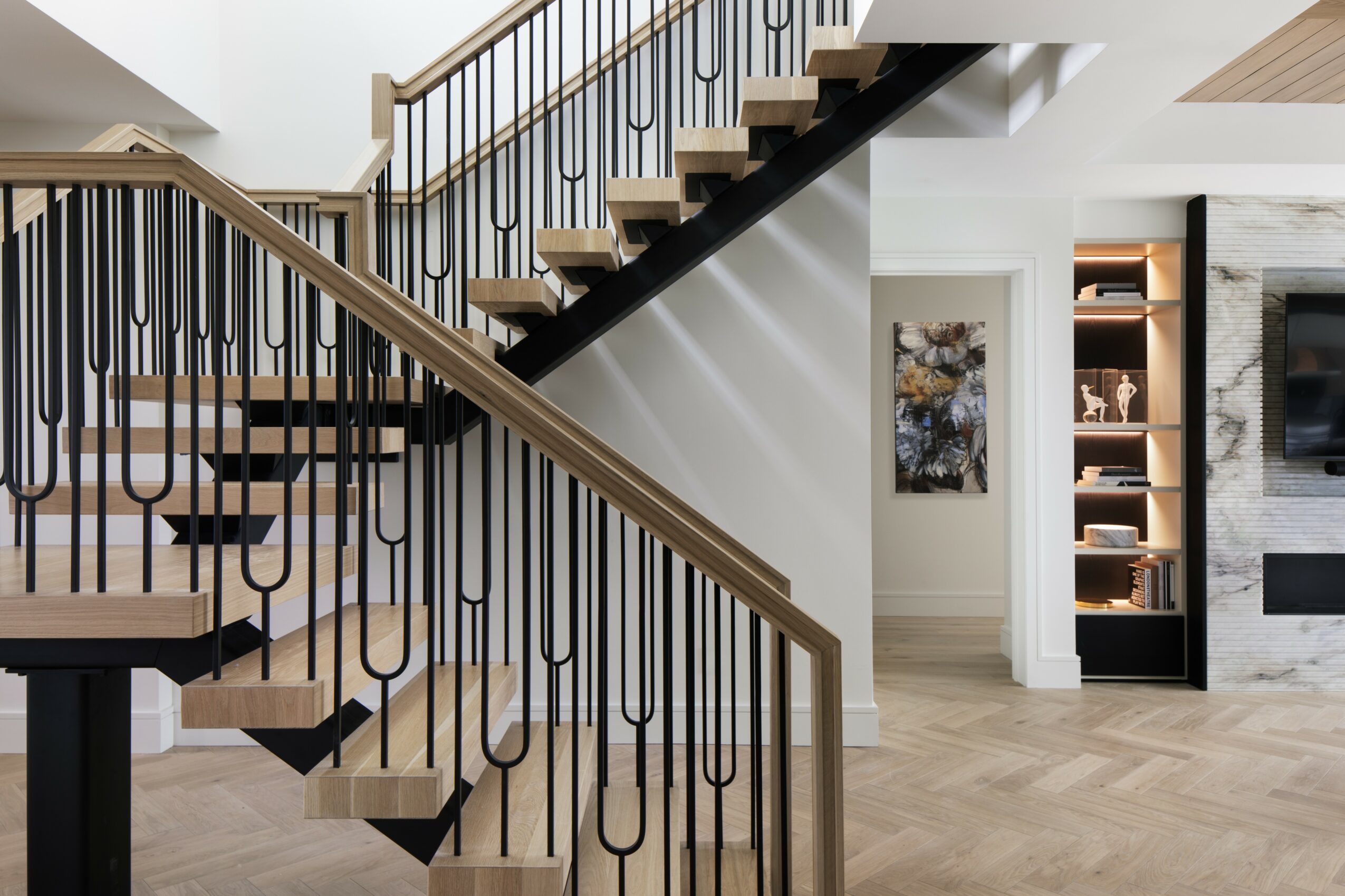 Interior staircase with wooden handrails and black metal balusters, leading to upper floor, with a living space in the background.