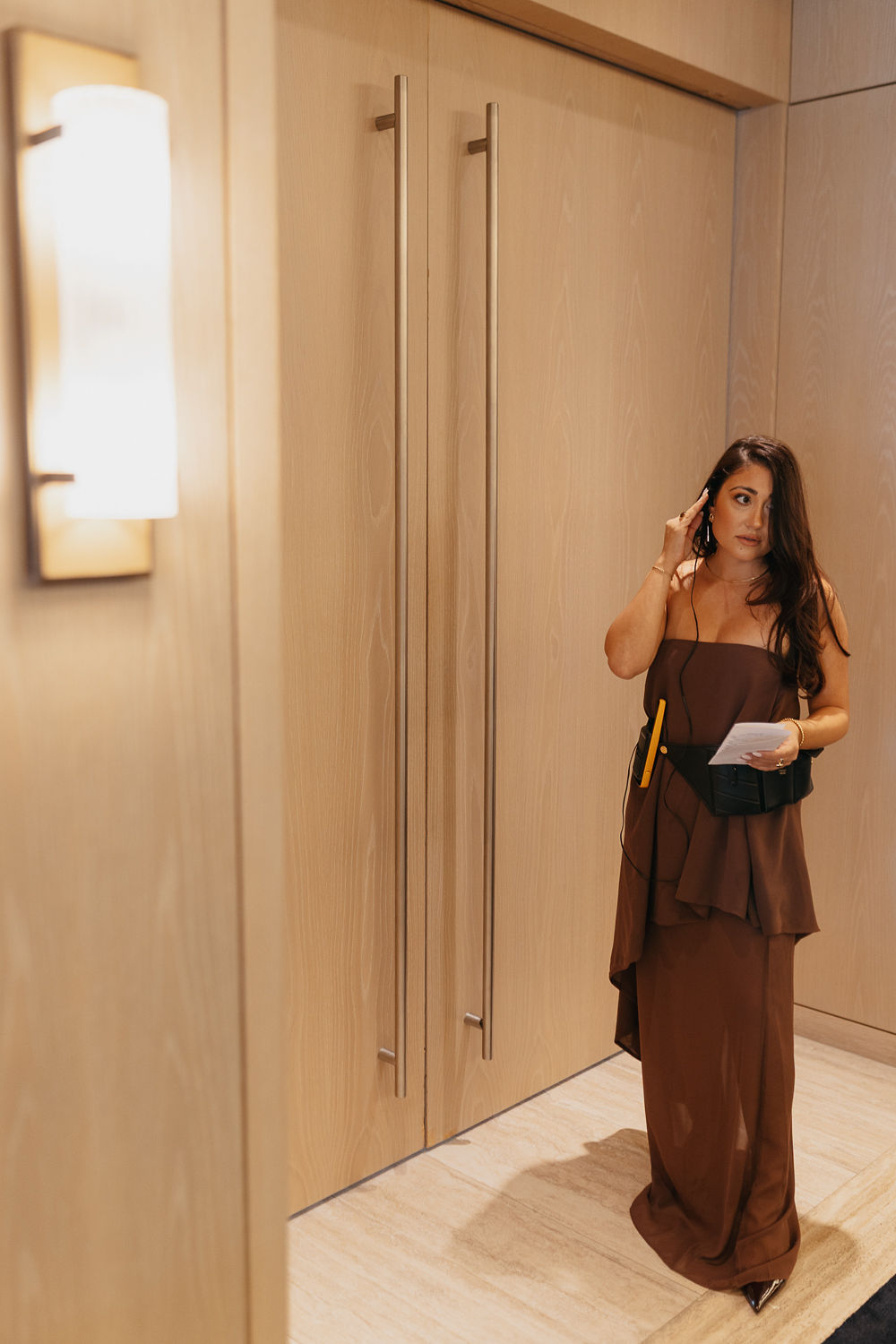Woman in a strapless dress holding papers, standing near a wooden door in an indoor setting.