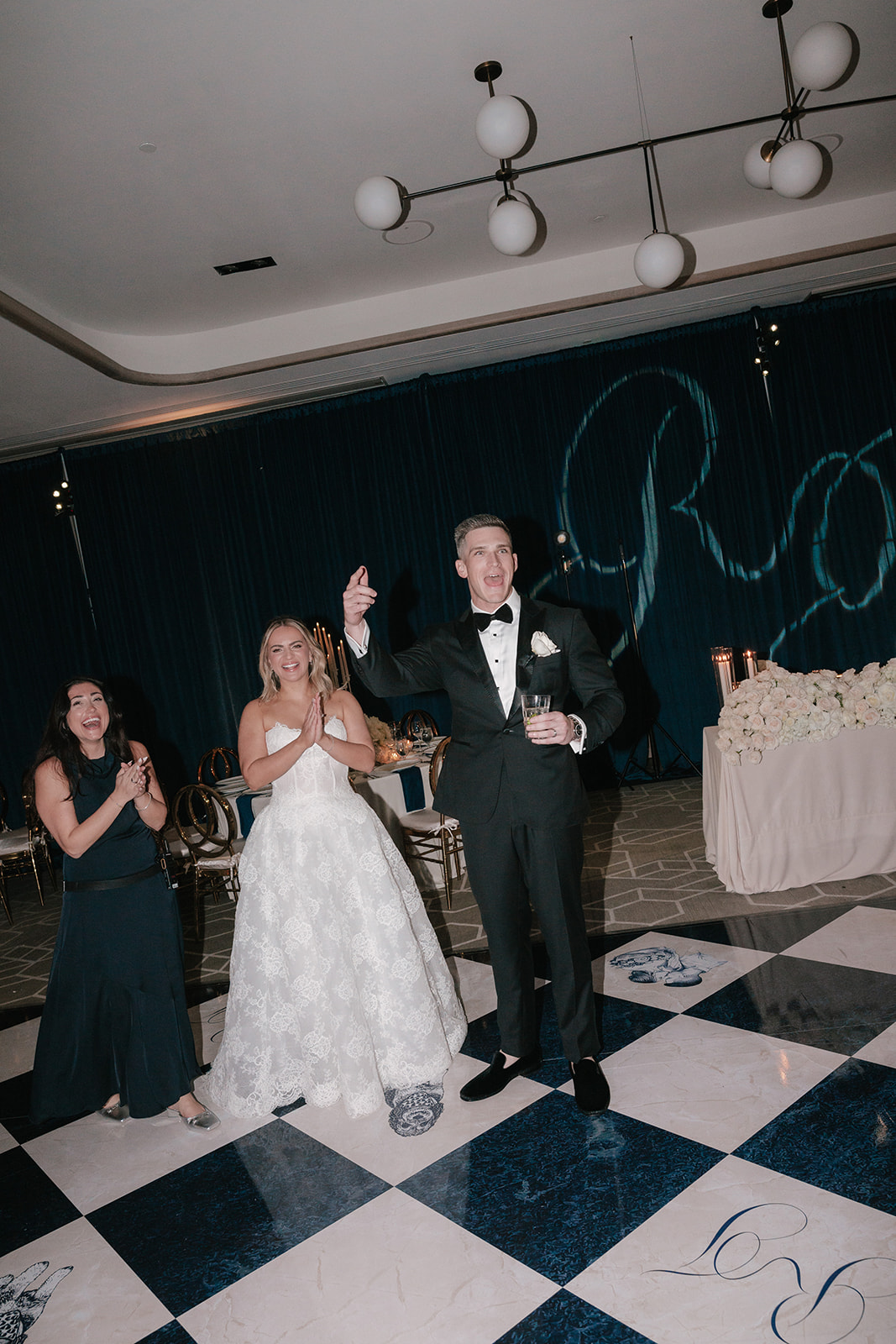 People celebrating at a formal event with a checkered floor and a dark backdrop with large letters, one person in a tuxedo, others clapping.