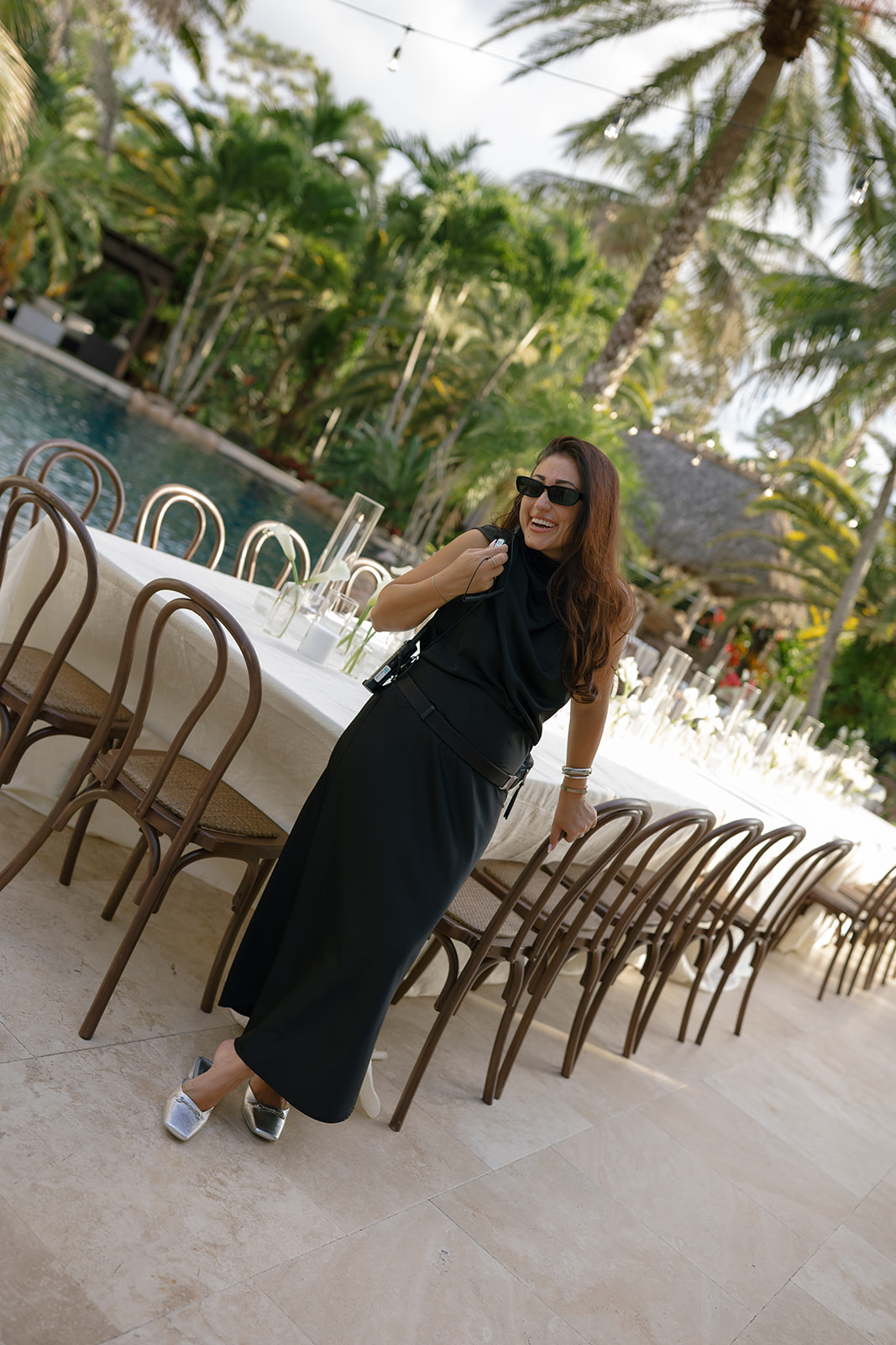 Woman in black dress and sunglasses standing outdoors near a long table with chairs, tropical trees in background.