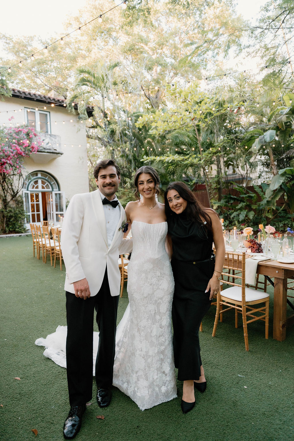 Three people standing outdoors, one in a wedding dress, two in formal attire, smiling, with trees and outdoor furniture in background.