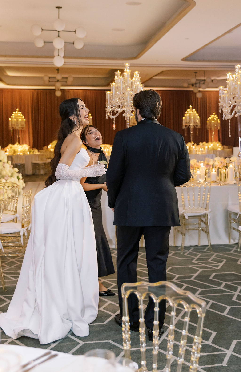 Three people in formal attire standing in a decorated banquet hall with chandeliers and tables, engaged in conversation.