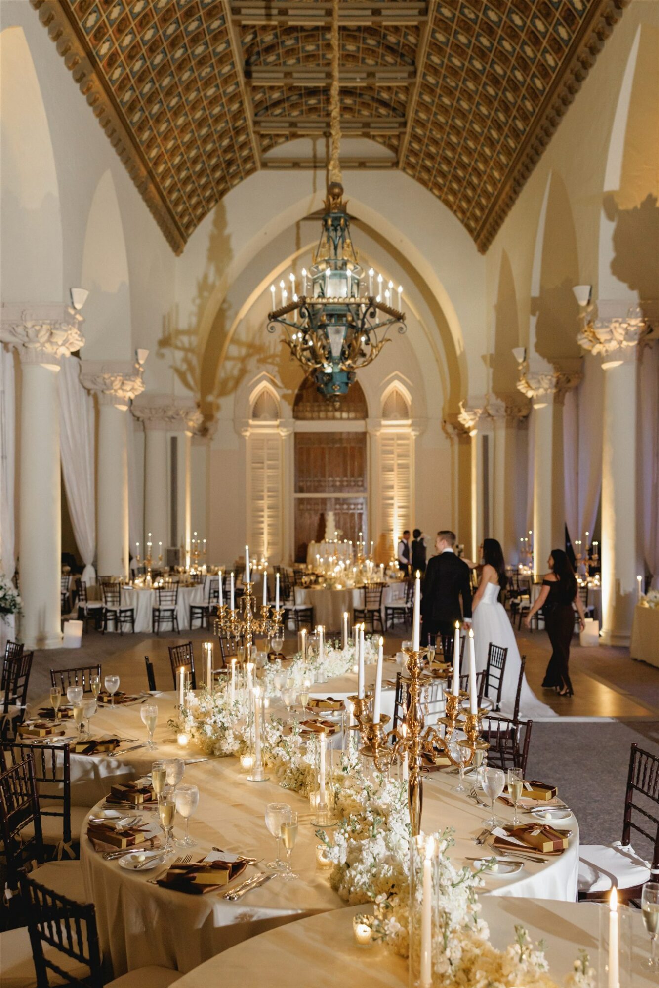 Elegant banquet hall with high arched ceiling, chandeliers, decorated tables, and people in formal attire.