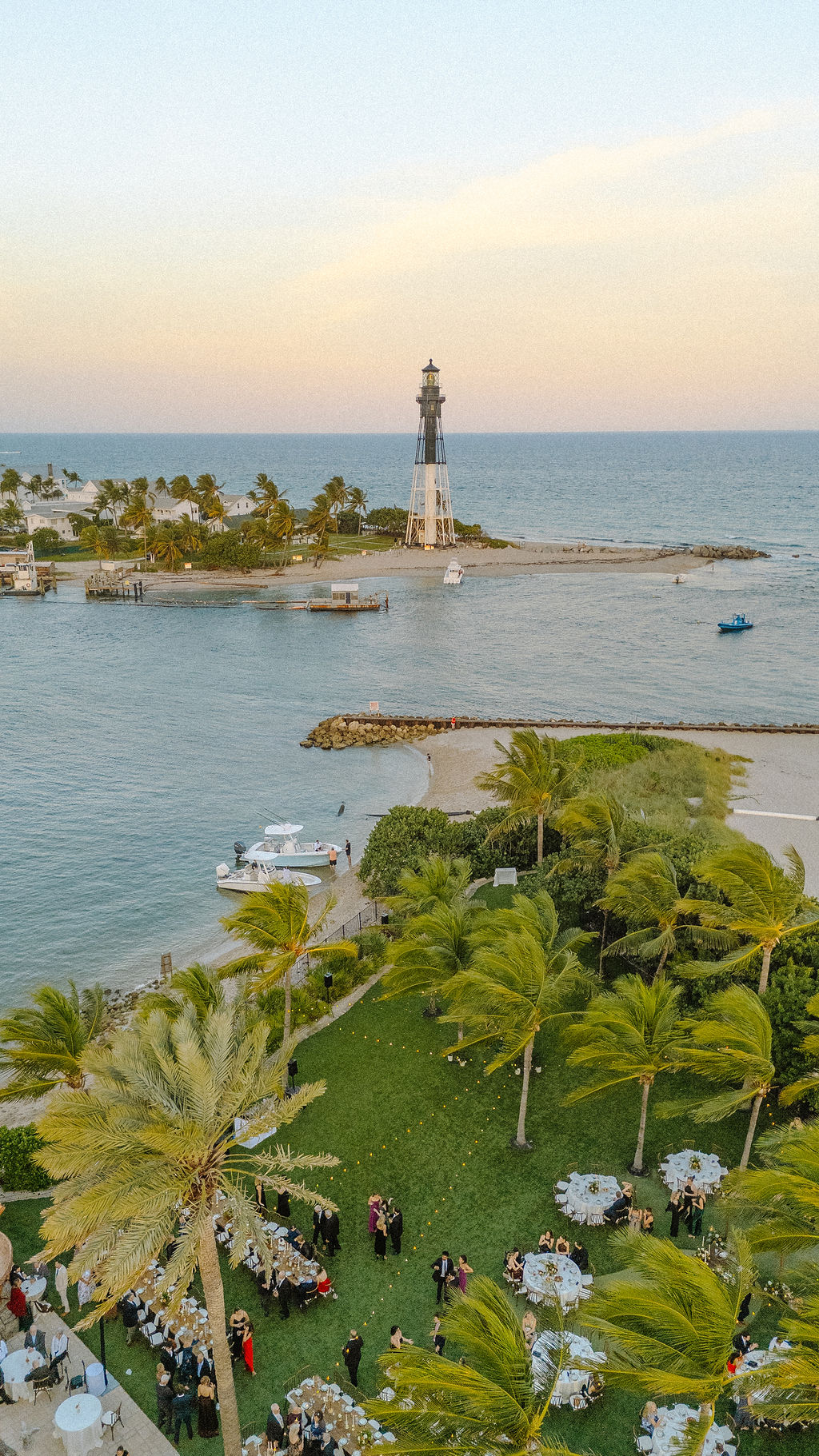 Lighthouse on a small island with palm trees, boats in water, and a green park with tables and people.