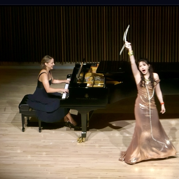 Two women perform on stage; one plays piano, the other sings with a feathered fan, wearing a long gown.