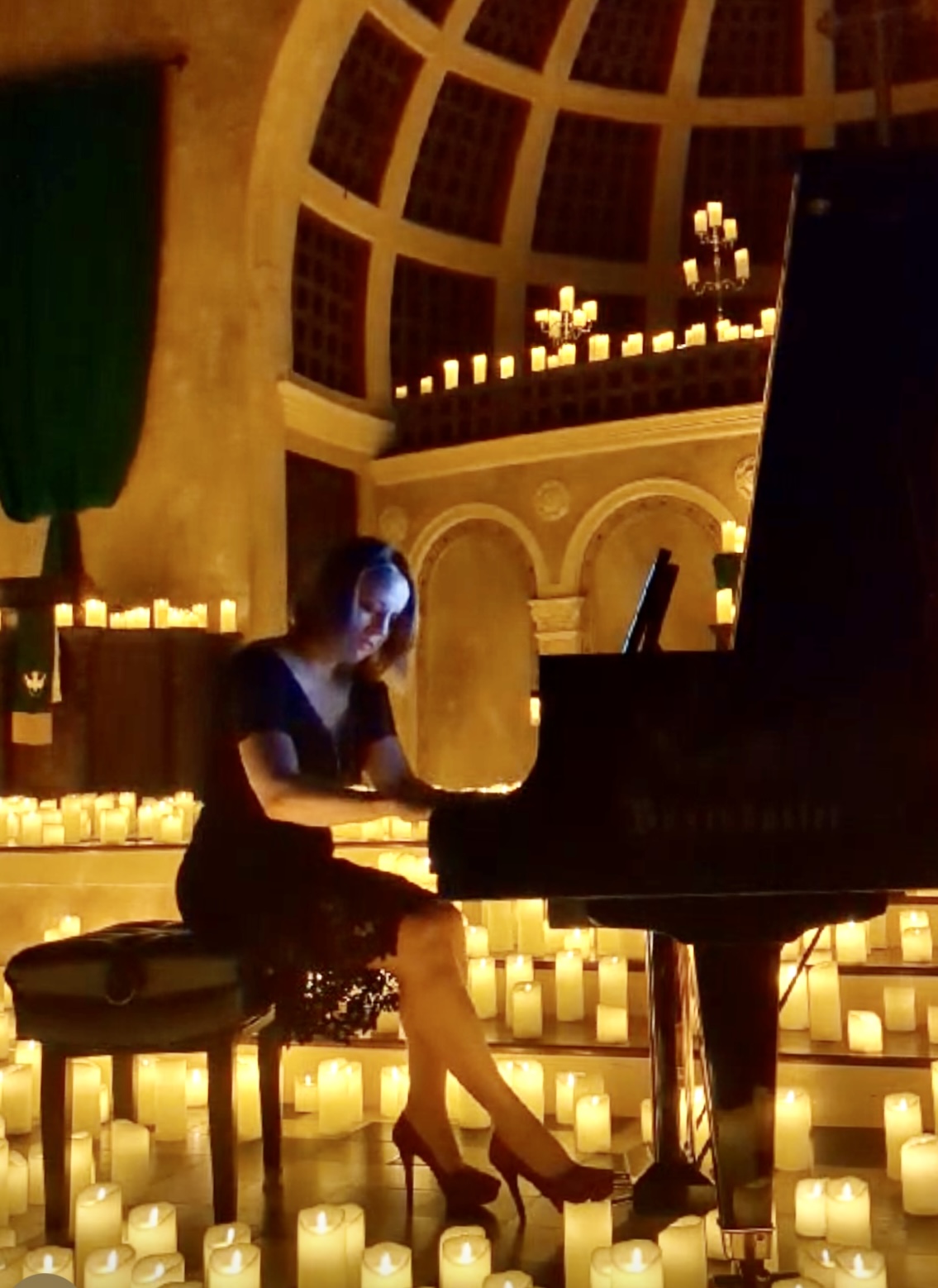Woman playing grand piano in a candlelit, domed room with candles on the floor and walls.