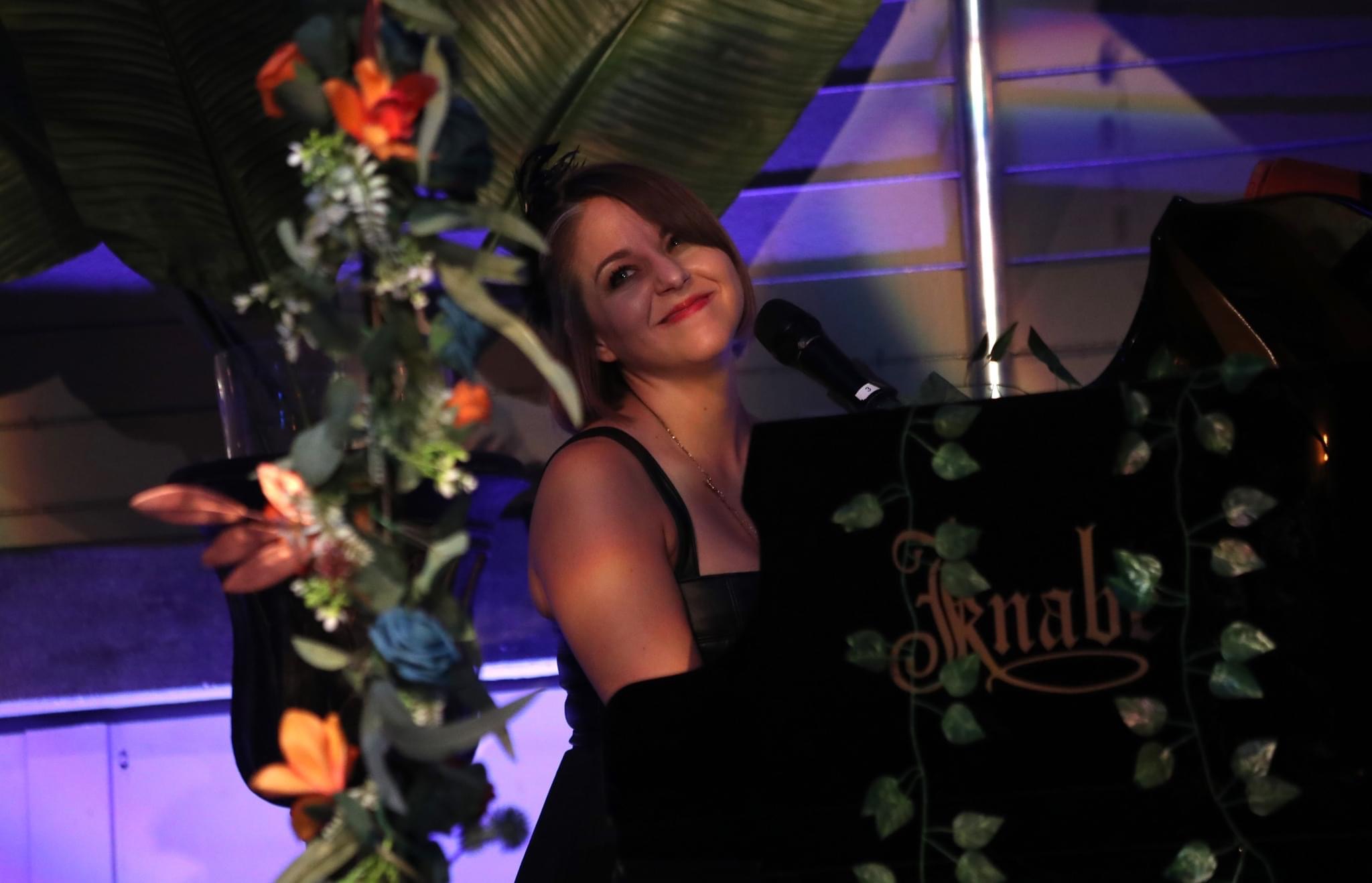 Woman with short hair smiling at a piano decorated with flowers, in a dimly lit setting.