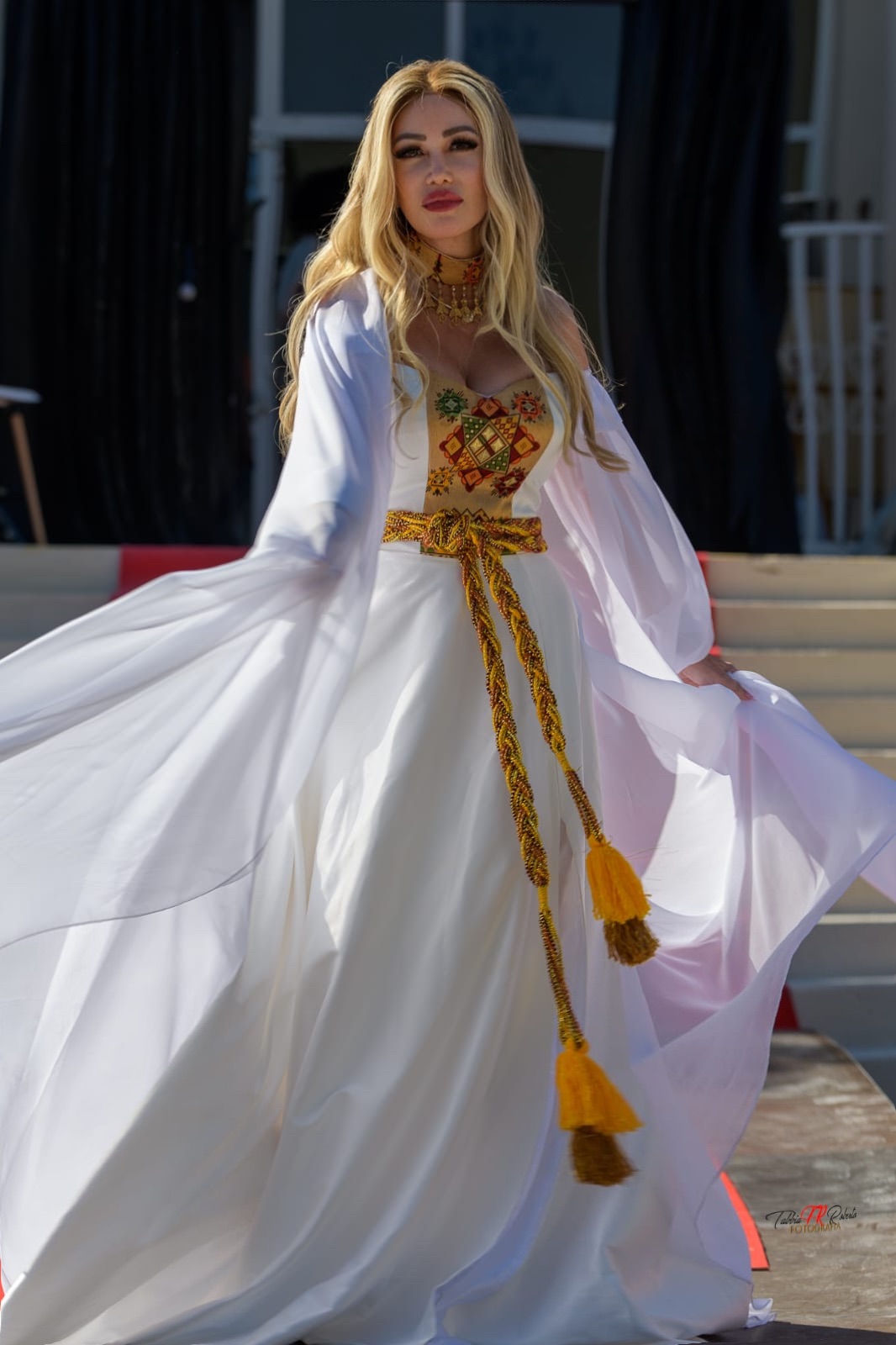 Woman in white dress with embroidered top and yellow tassel belt, standing outdoors with flowing fabric.