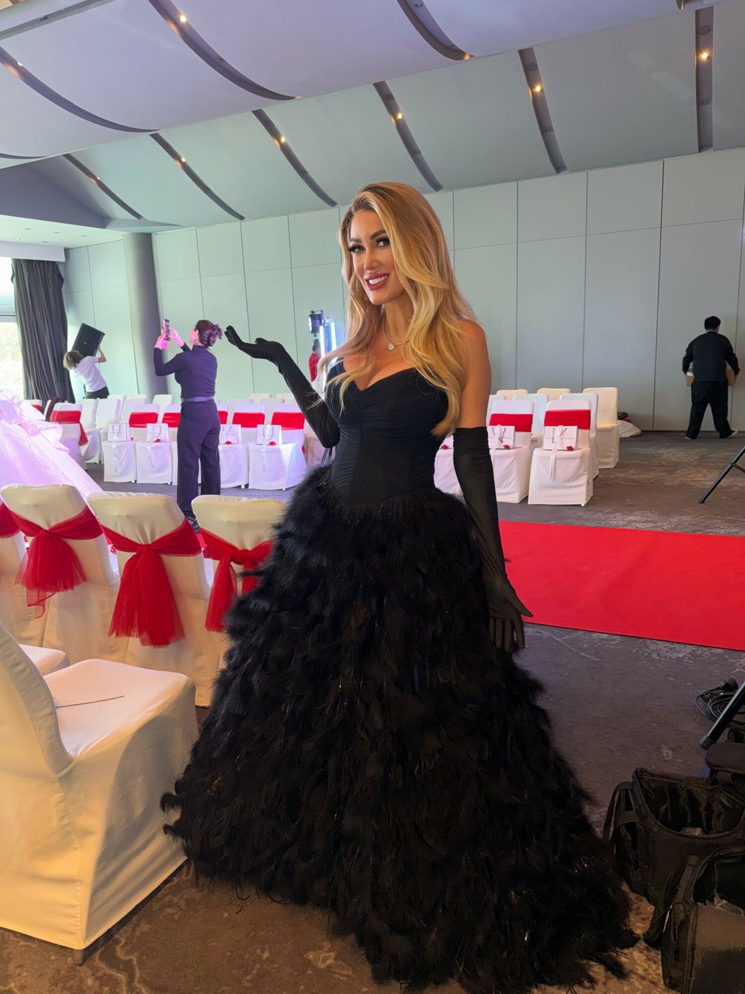 Woman in a black gown with long gloves, smiling at an event with decorated chairs and a red carpet.