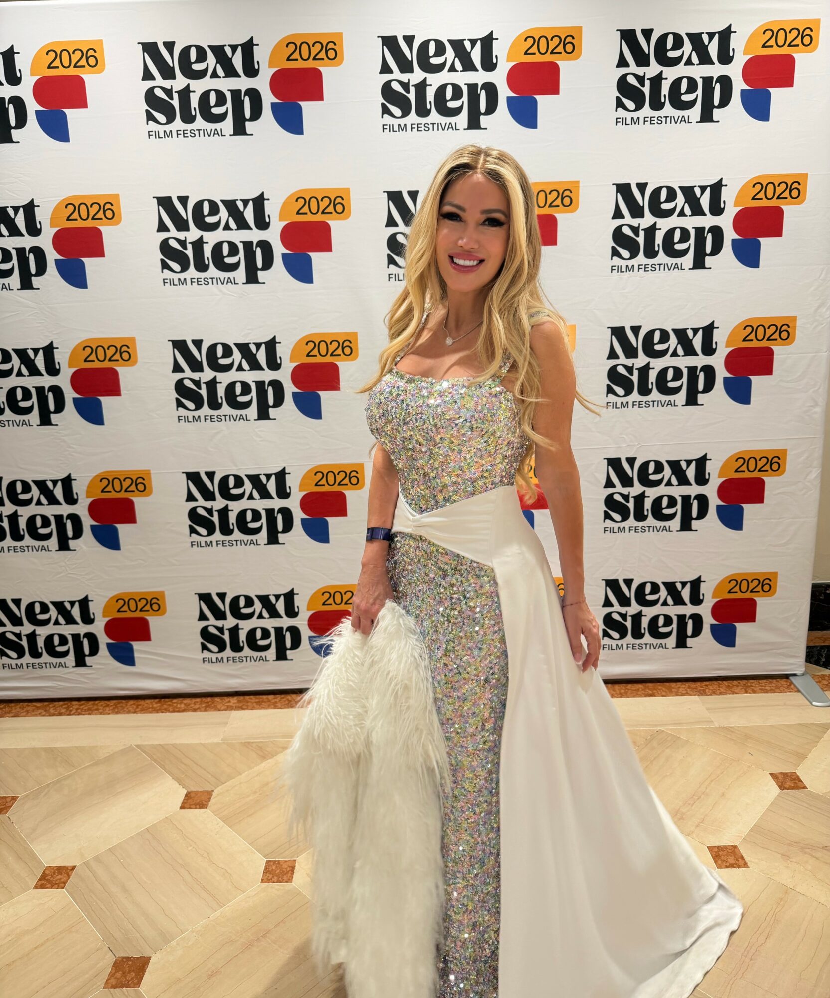Young woman in a sparkling gown with a fitted bodice and flowing skirt, smiling at the camera, standing in front of a step-and-repeat backdrop.