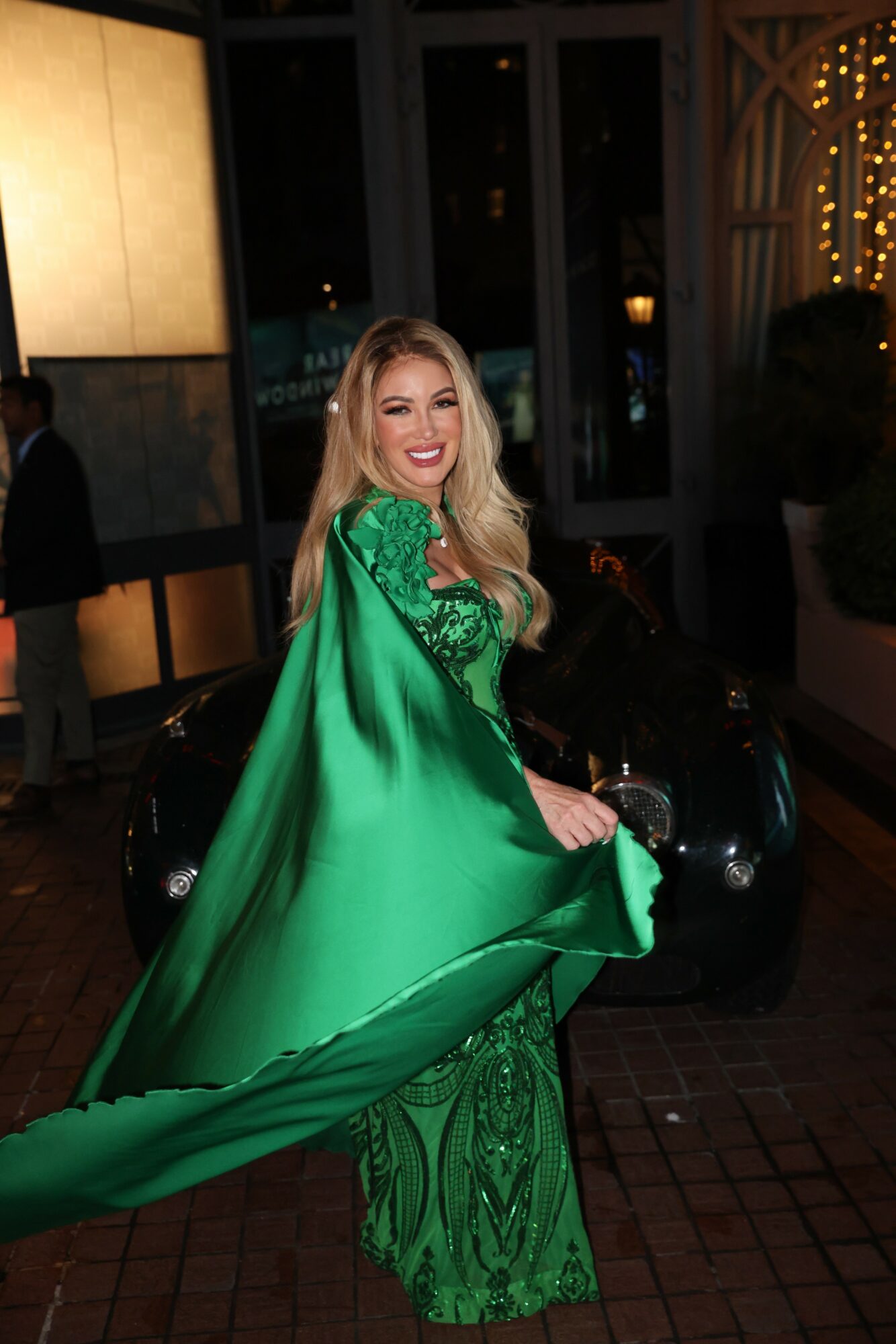 Woman in green dress smiling, holding the dress out, nighttime setting with lights and a car in background.