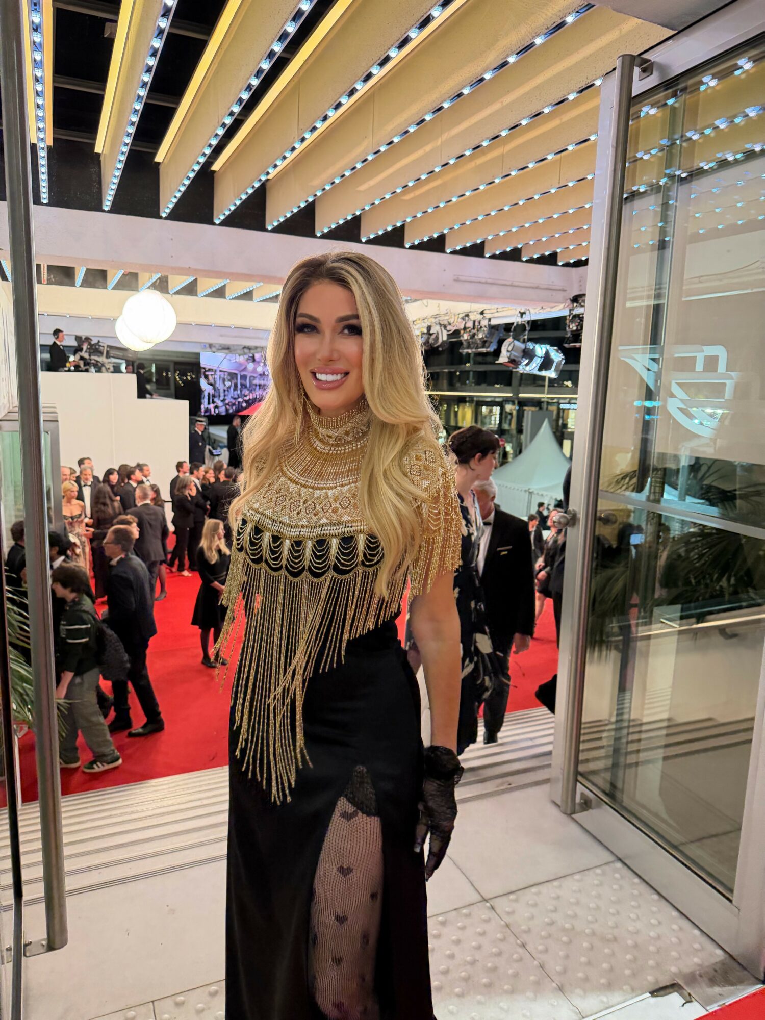 Woman with long blonde hair in a black dress with gold fringe, smiling at an event with a crowd and red carpet.