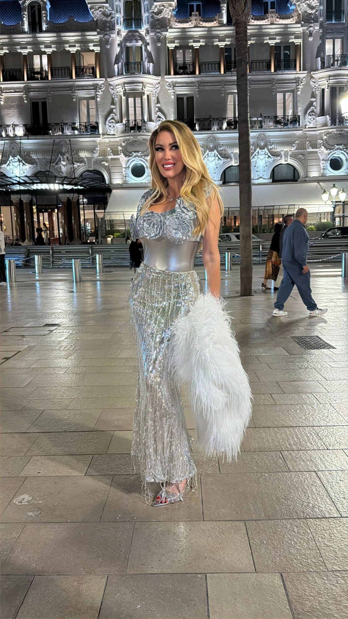 Woman in a silver and white gown holding a white fur stole, standing on a city street at night.