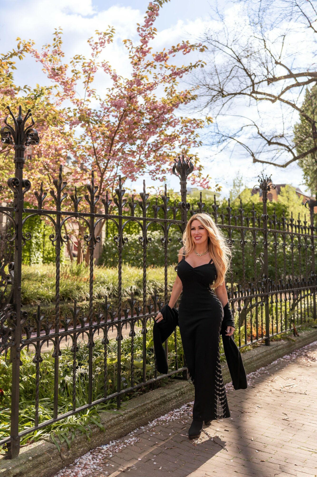 Woman in black dress standing by a black iron fence with trees and pink blossoms in background.
