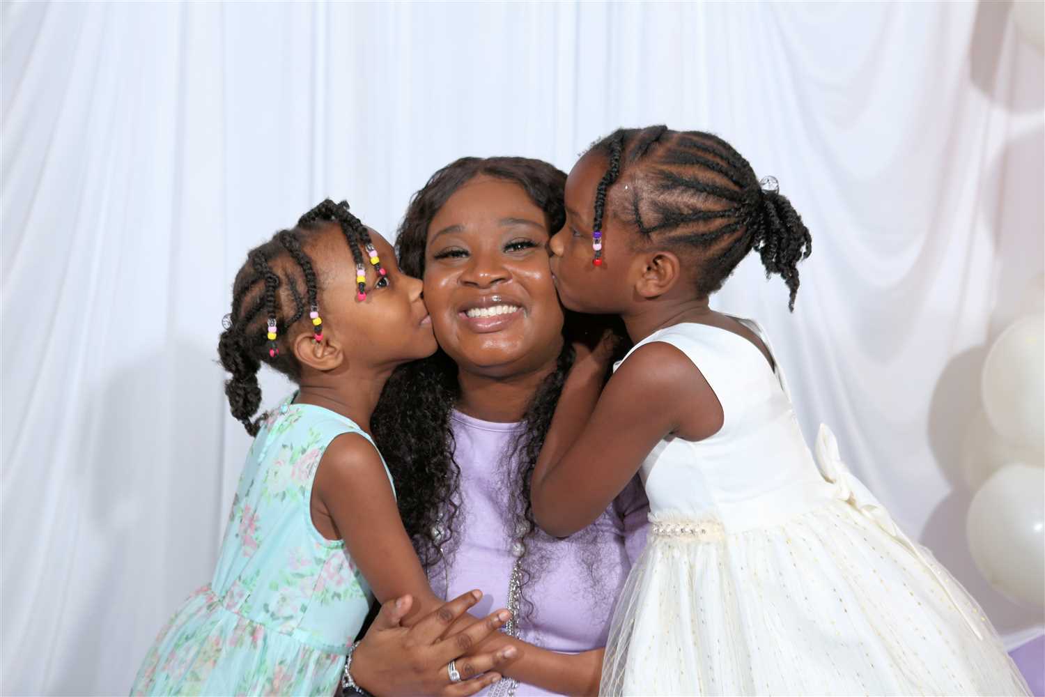 Two children kiss a smiling woman on each cheek, with a white backdrop and balloons nearby.