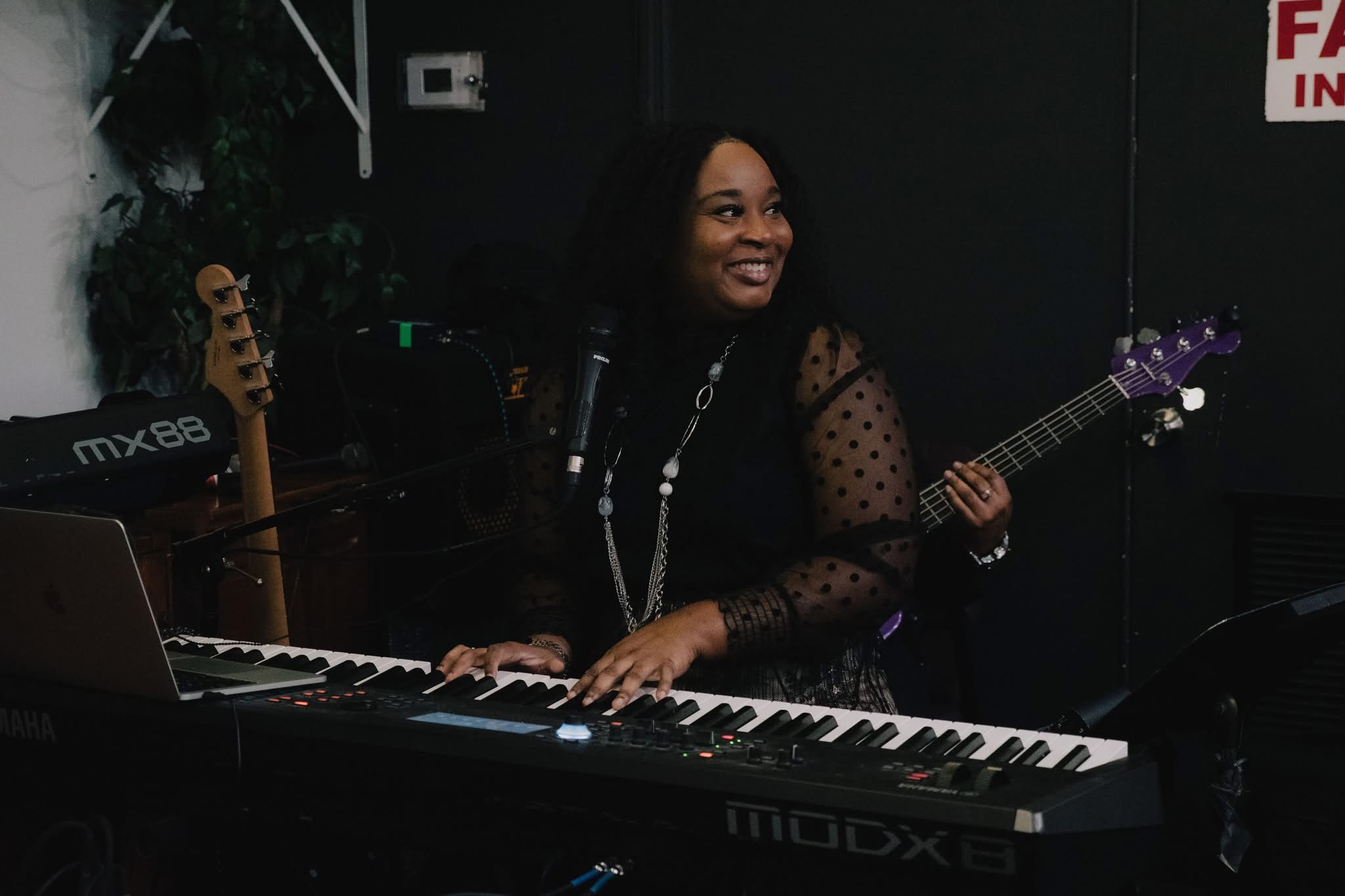 Woman playing keyboard and holding guitar, smiling, in a dark room with music equipment.