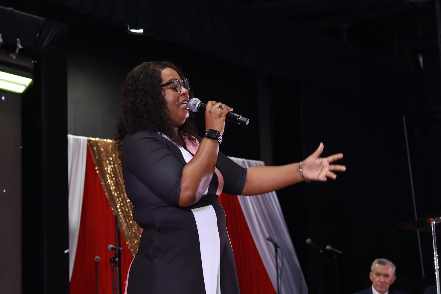 Woman with curly hair and glasses speaking into a microphone on stage, gesturing with her right hand, with red and white drapes behind her.
